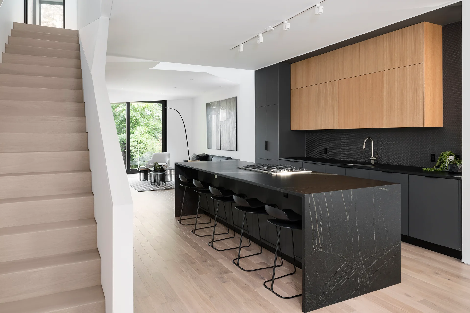 A minimalist kitchen with a black stone waterfall island, two-tone wood and black cabinets, and an adjacent light wood staircase.