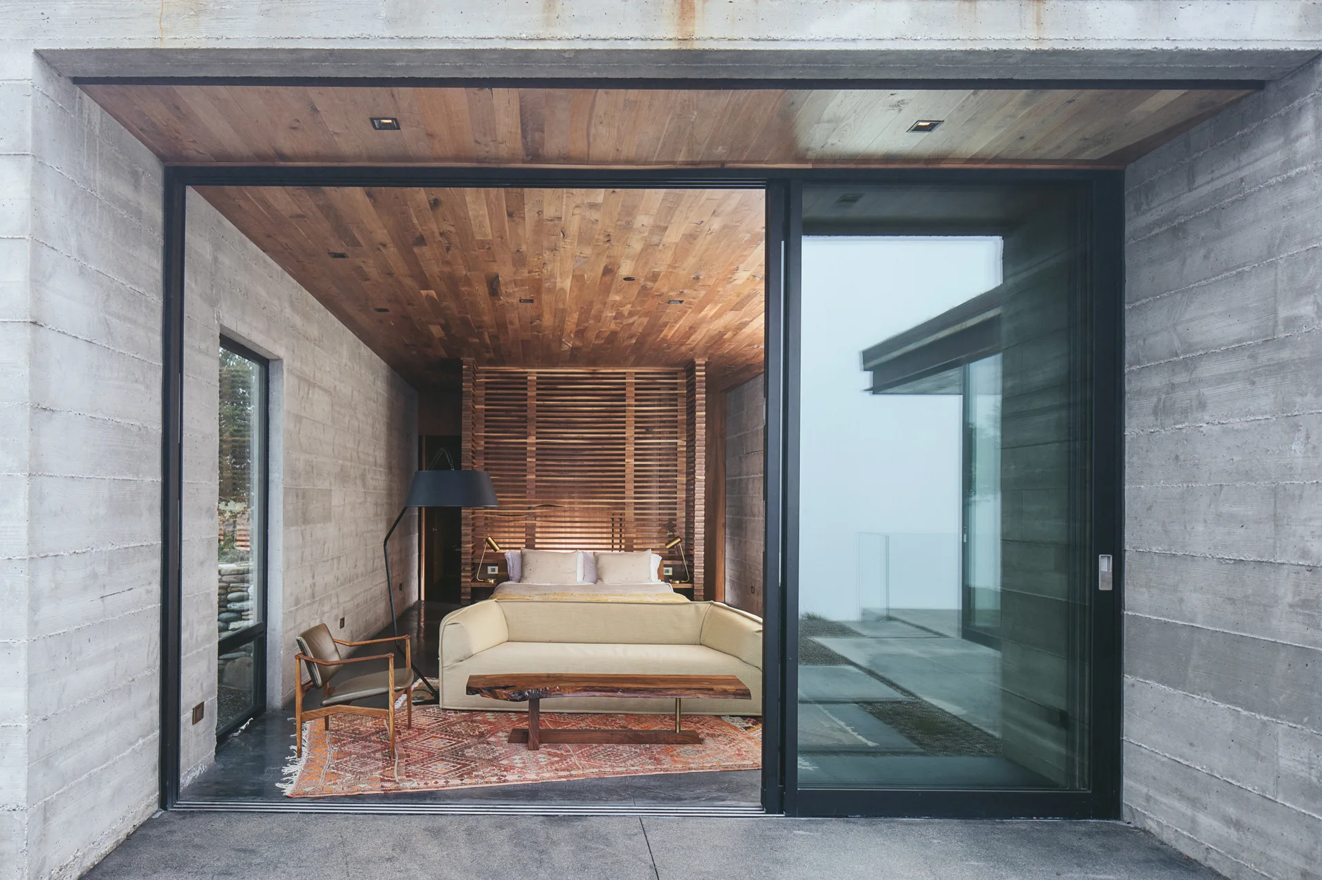 A modern bedroom featuring board-formed concrete walls, a wood plank ceiling, a cream sofa, and a large sliding glass door.