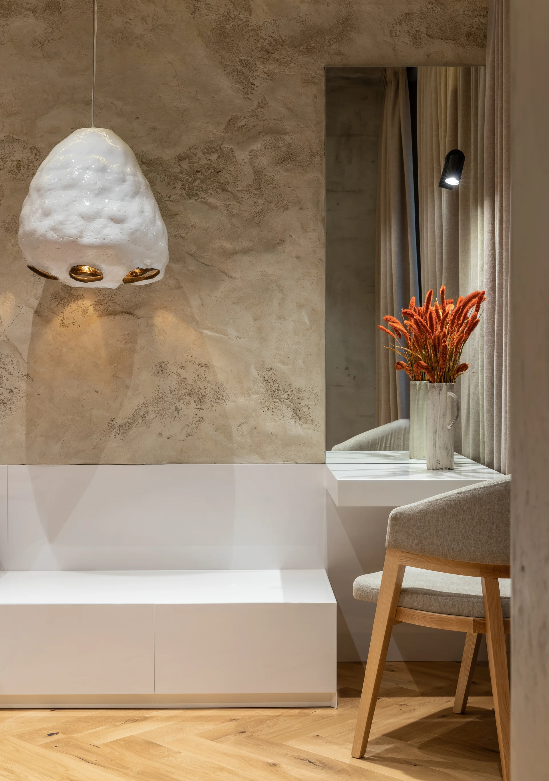 A modern dressing vanity with a white console, a wooden chair, an organic white pendant light, and a rough textured beige plaster wall.