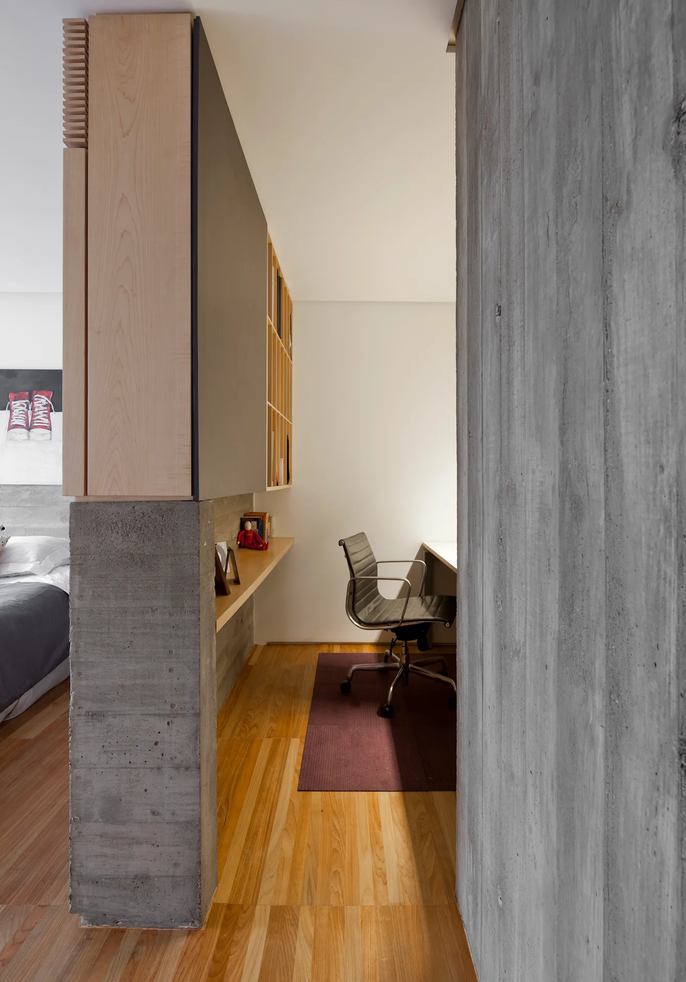 A modern home office nook features a built-in wooden desk, open shelving, a black desk chair, and a textured concrete partition wall.