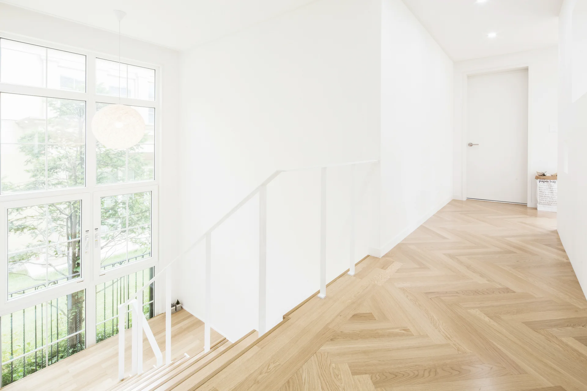 A bright hallway with light wood herringbone flooring features a white staircase, a tall multi-pane window, and white walls.
