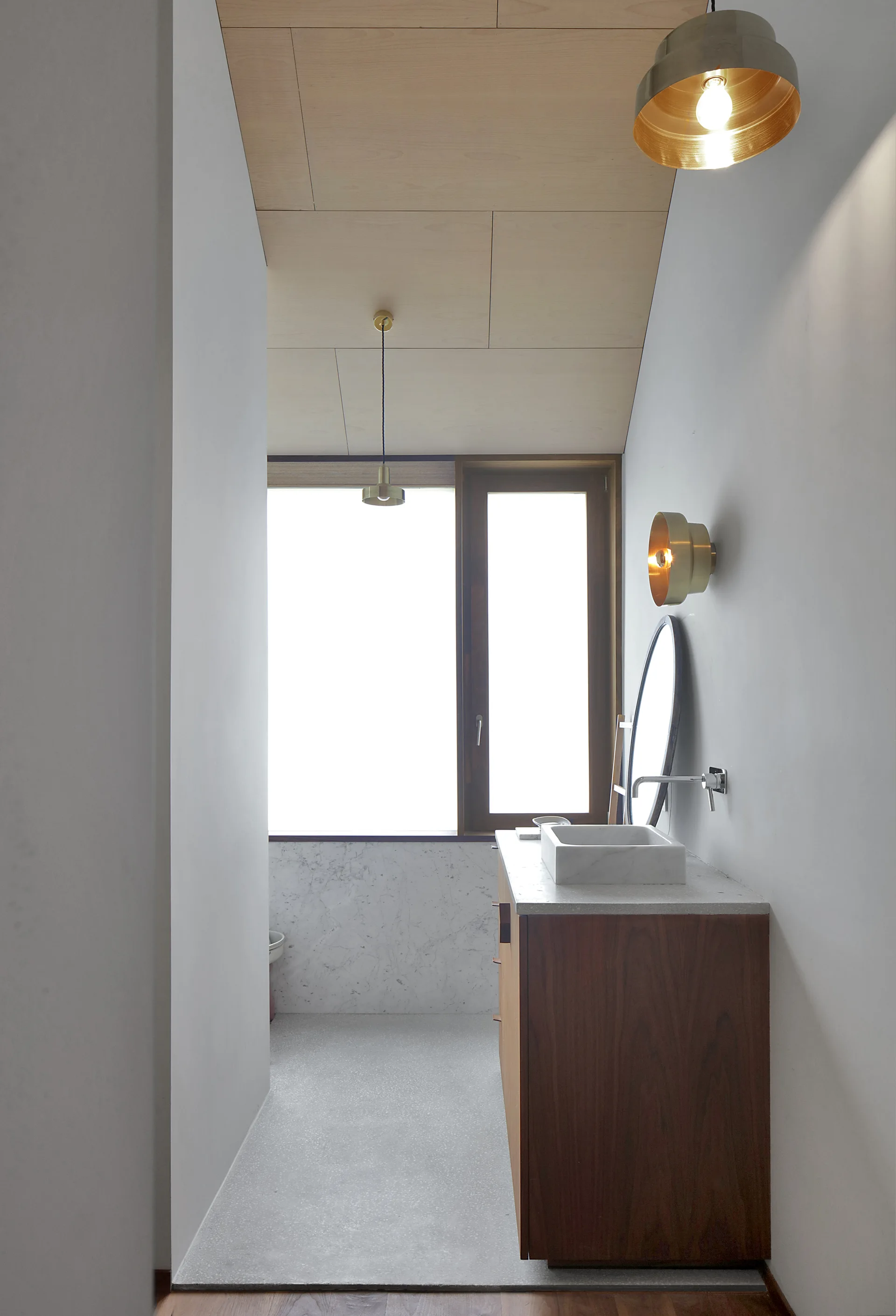 A narrow bathroom features a walnut vanity with a stone sink, a round wall-mounted mirror, brass light fixtures, and a light wood paneled ceiling.