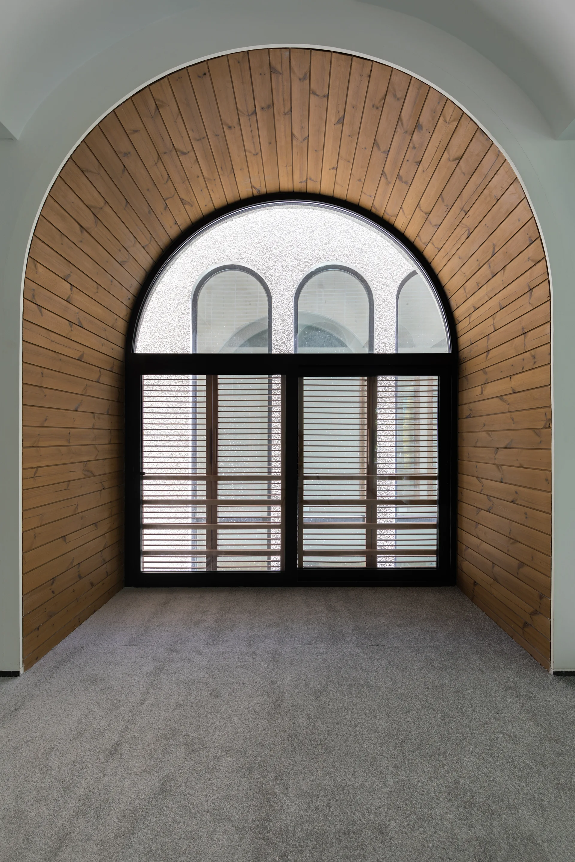 A large arched window with horizontal blinds is set into a deep alcove lined with warm wood paneling above gray carpet.