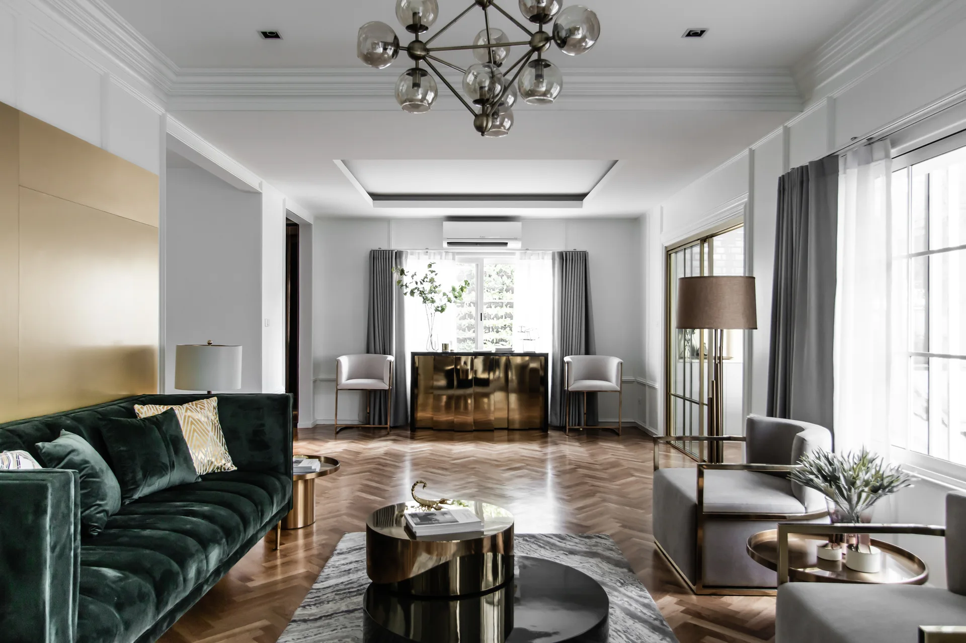 A modern living room features an emerald green velvet sofa, grey armchairs, gold coffee tables, white paneled walls, and herringbone wood floors.