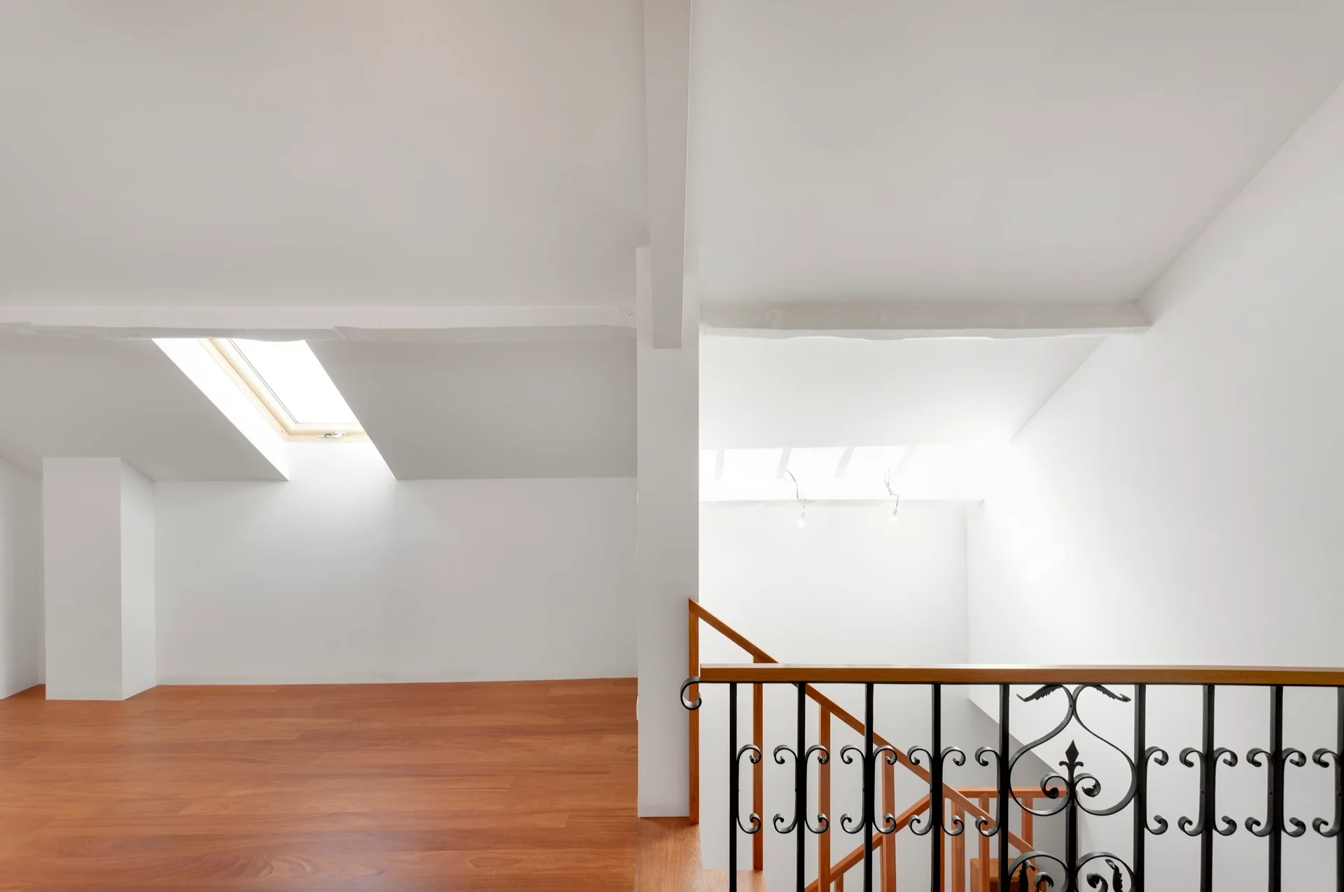 A bright, empty attic space features warm wood floors, two skylights, and a staircase with an ornate black wrought-iron railing.