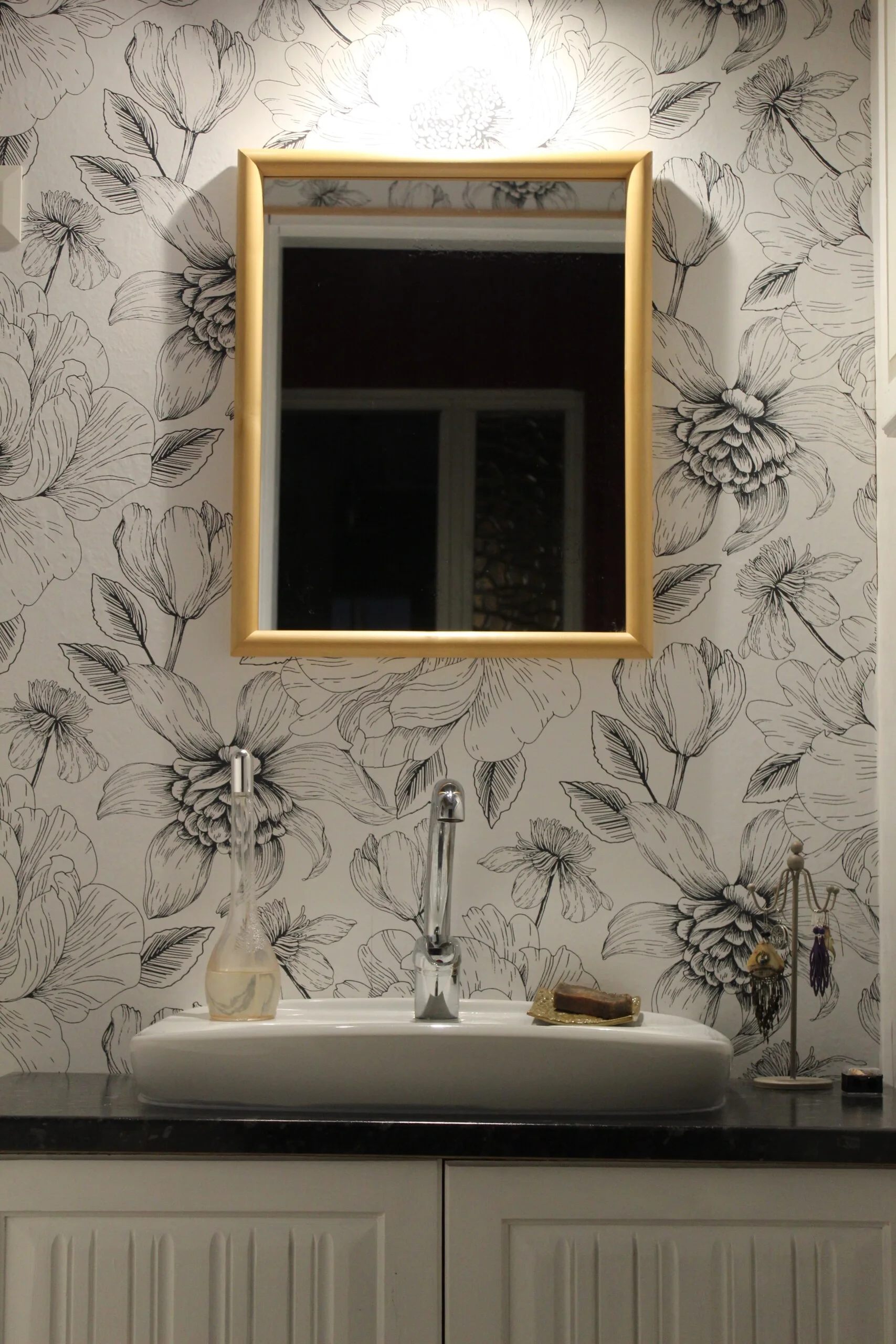 A bathroom vanity features a white ceramic vessel sink, a gold-framed mirror, and a wall covered in black and white sketch floral wallpaper.