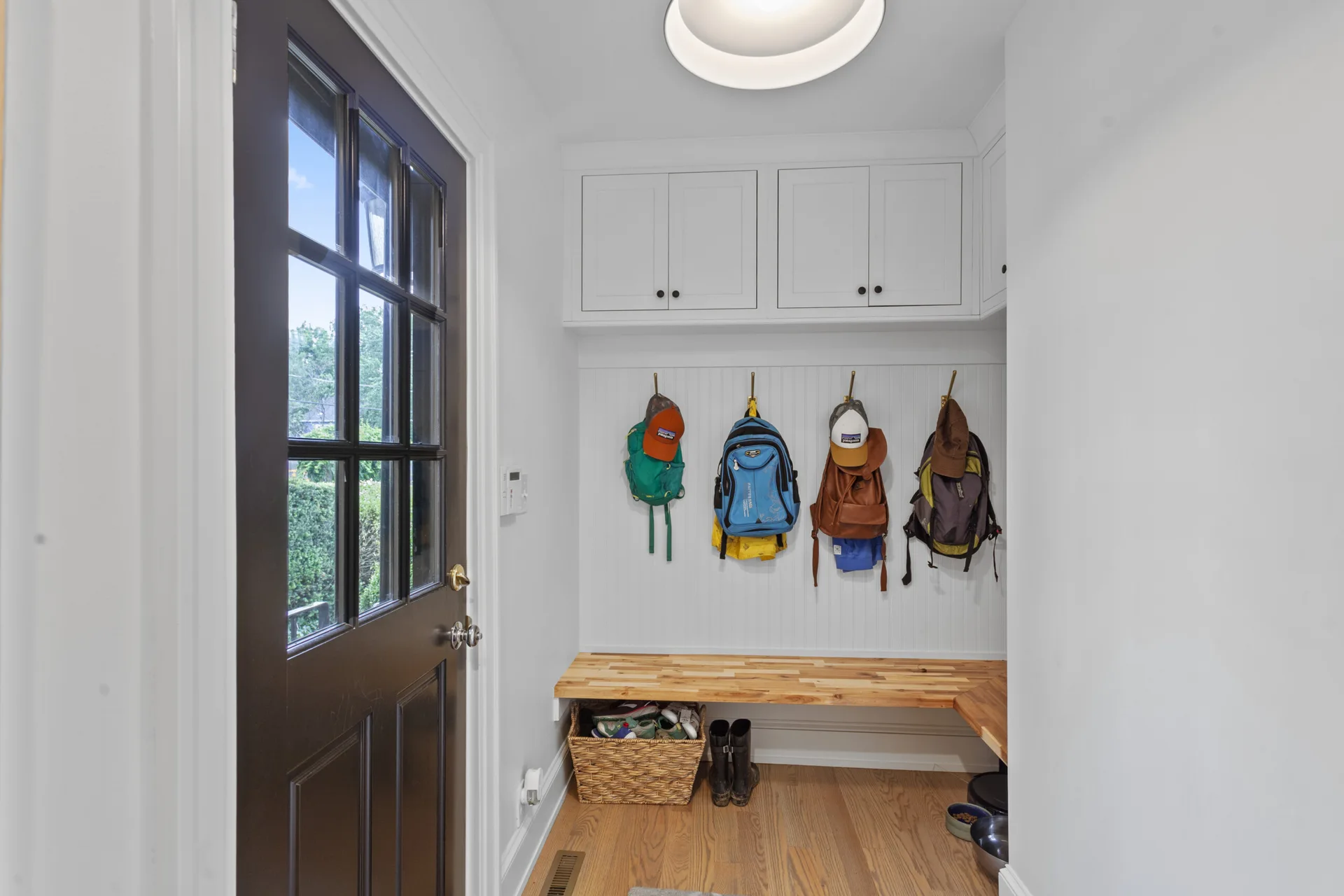 A white mudroom featuring an L-shaped wood bench, white upper cabinets, and gold hooks holding backpacks against a beadboard wall.