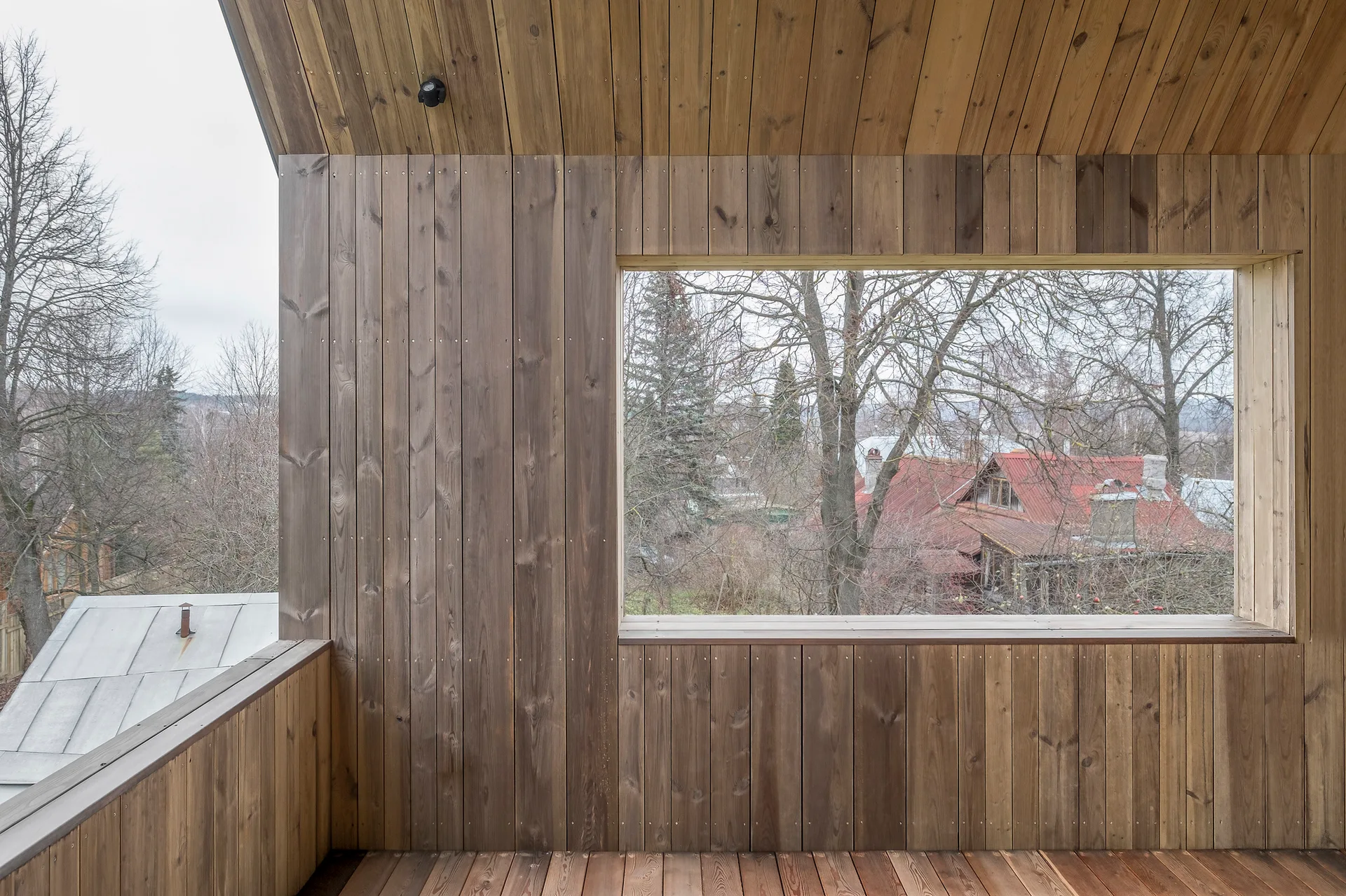 A covered wooden balcony clad in vertical timber planks features a vaulted ceiling and a large rectangular window overlooking trees and red rooftops.