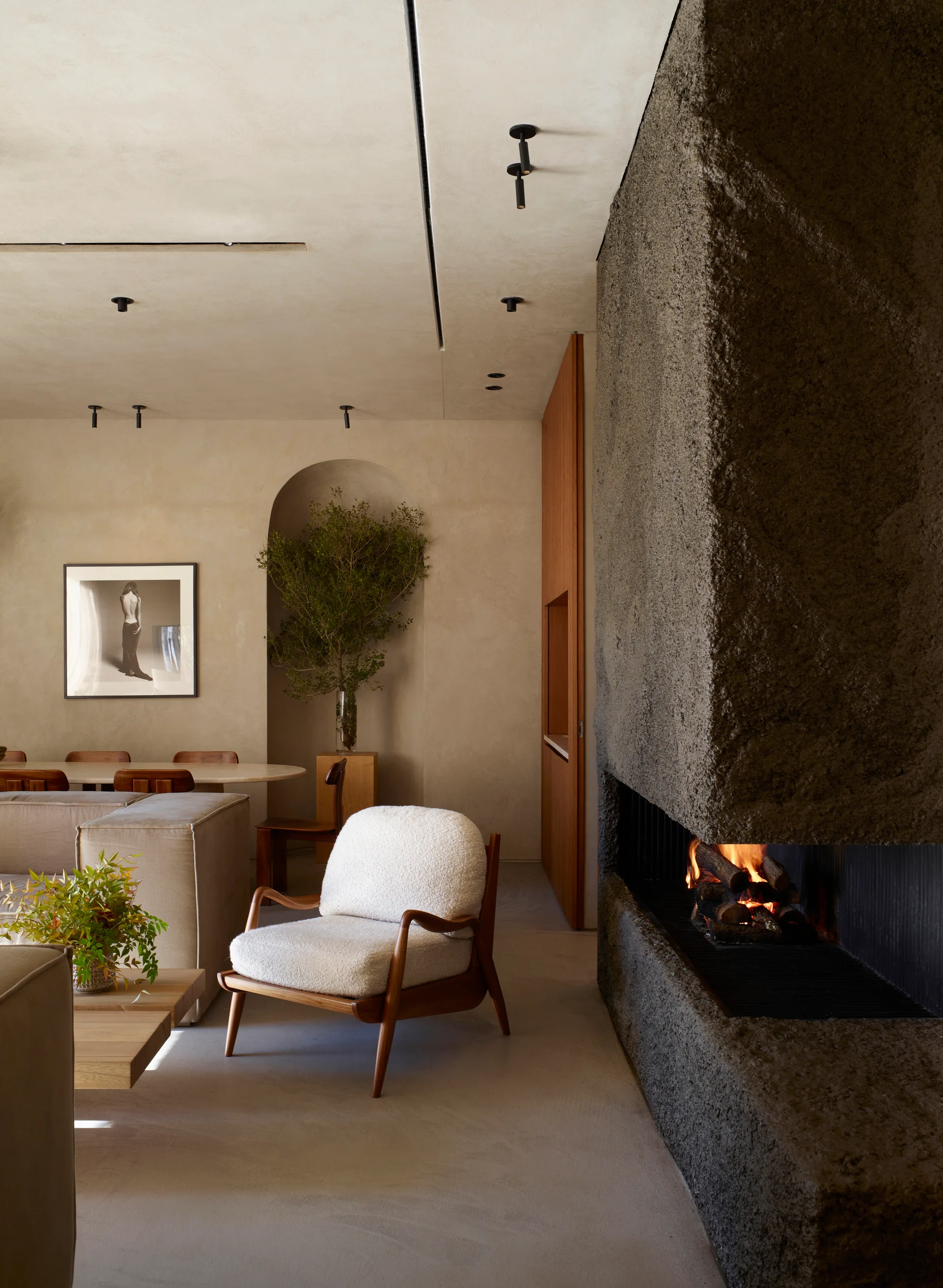 A living room features a textured dark stone fireplace alongside a white bouclé armchair and a recessed arched wall alcove holding a green plant.