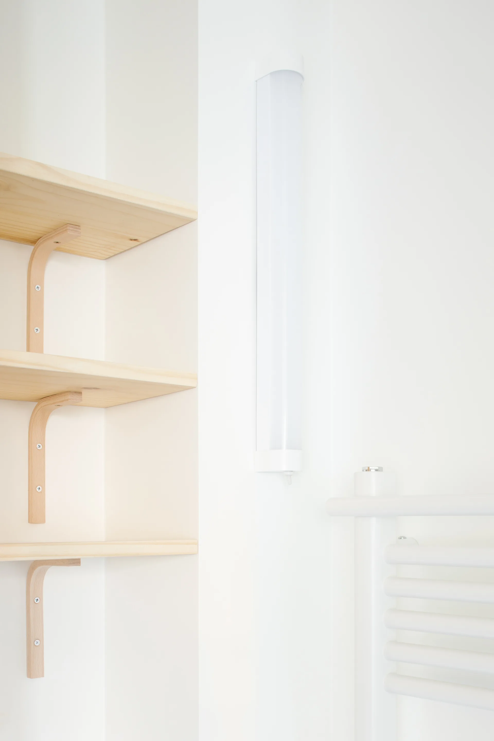 Three light wood shelves with matching wooden brackets mount to a white wall beside a vertical white linear light fixture.
