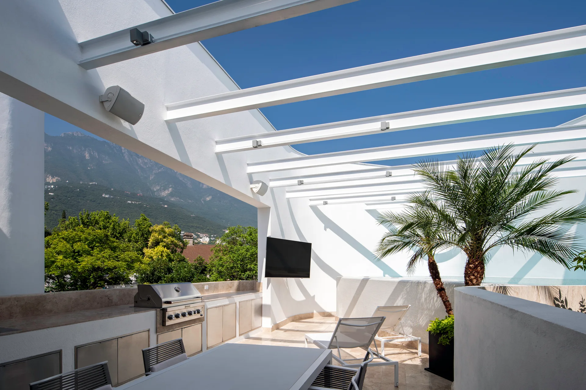 An outdoor kitchen with a built-in grill and dining table sits on a white tile terrace beneath pergola beams with a mountain background.