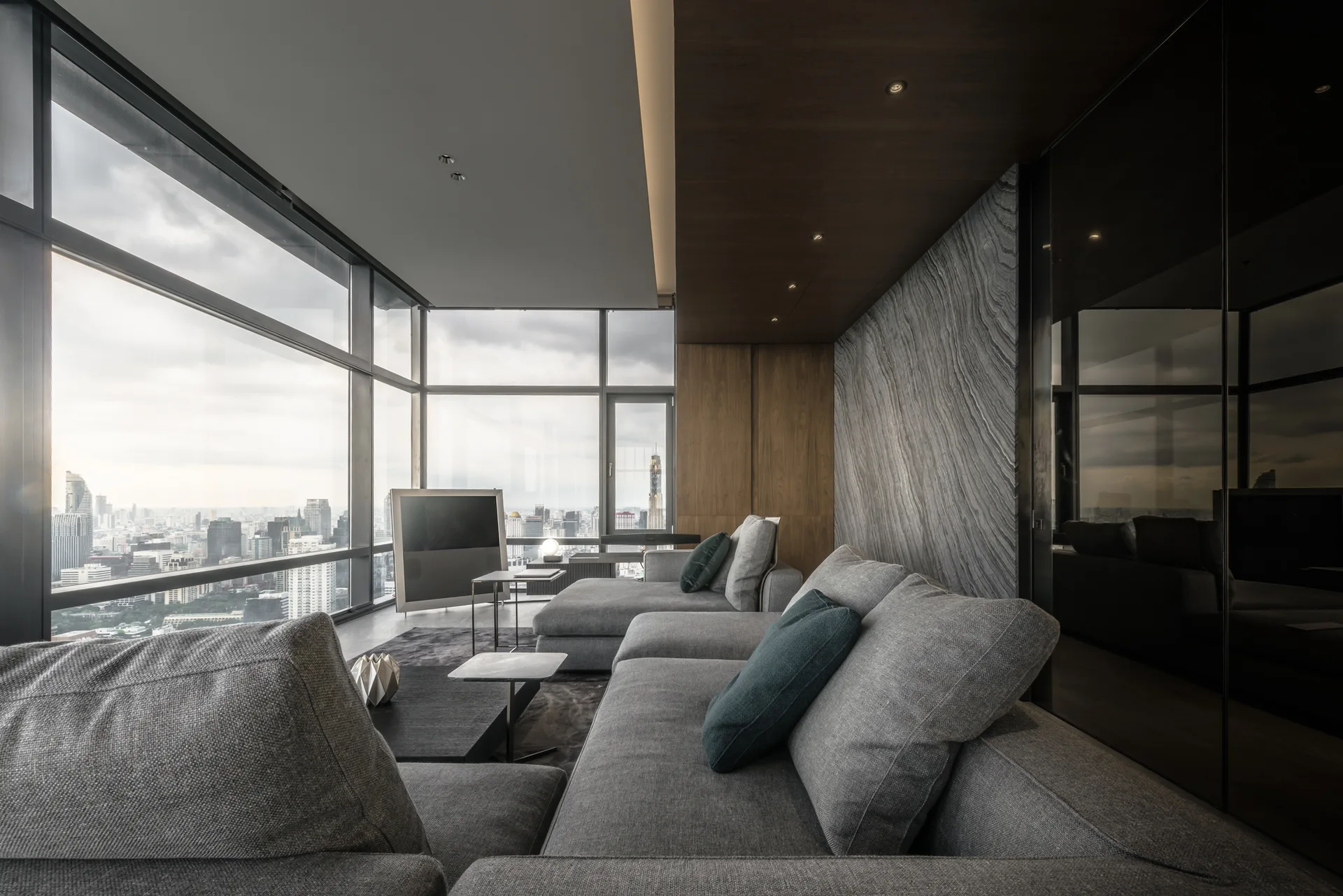 A modern living room features a large grey sectional sofa, textured grey stone wall paneling, a dark wood ceiling, and expansive city-facing windows.
