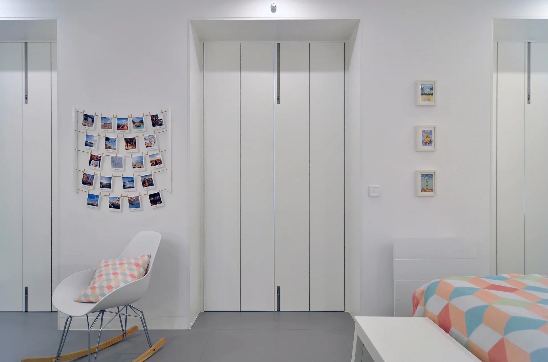 A minimalist white bedroom featuring tall folding closet doors, a white molded rocking chair with a geometric pillow, and a grid photo display.