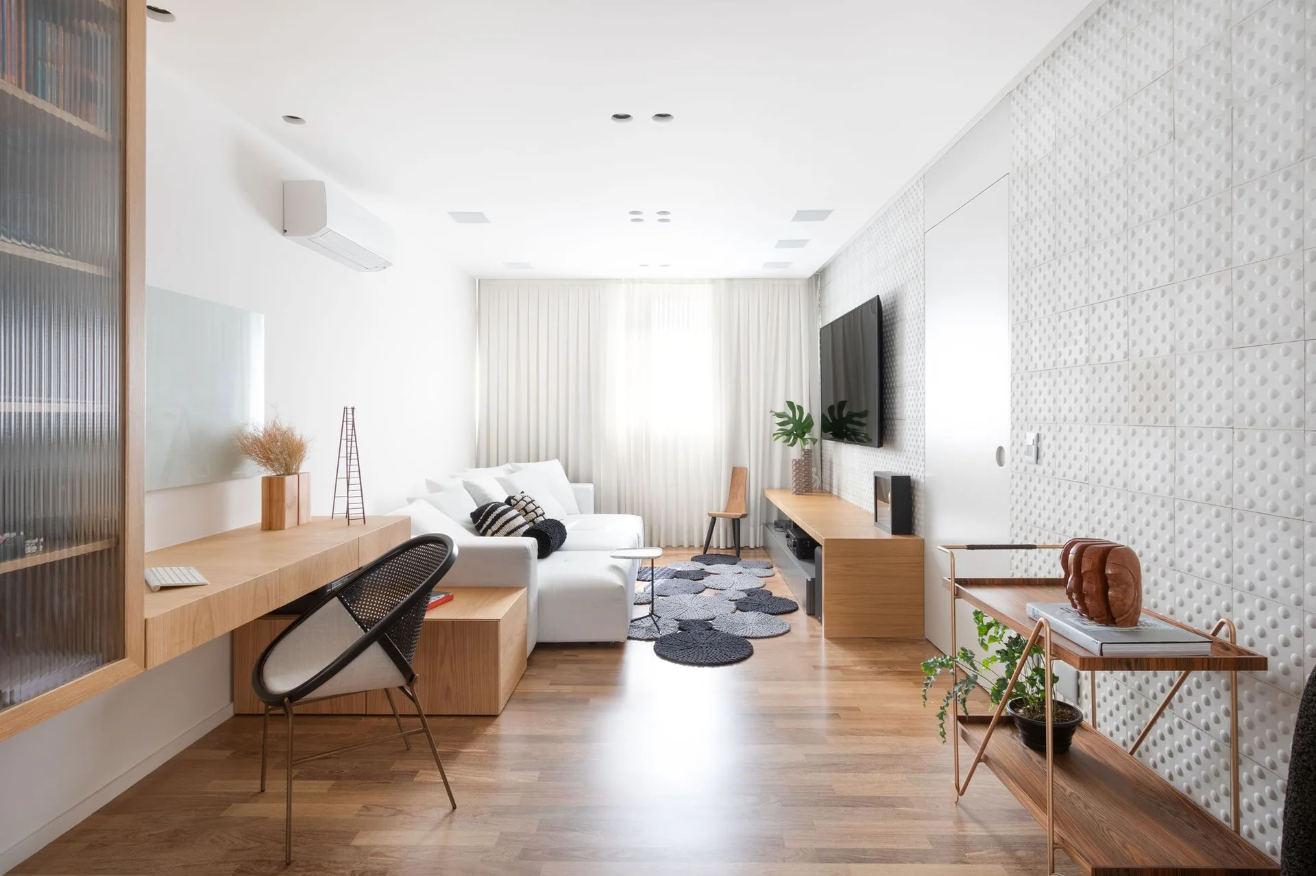 A long white living room features a sectional sofa, a floating wooden desk, a mounted television, and a white textured tile accent wall.