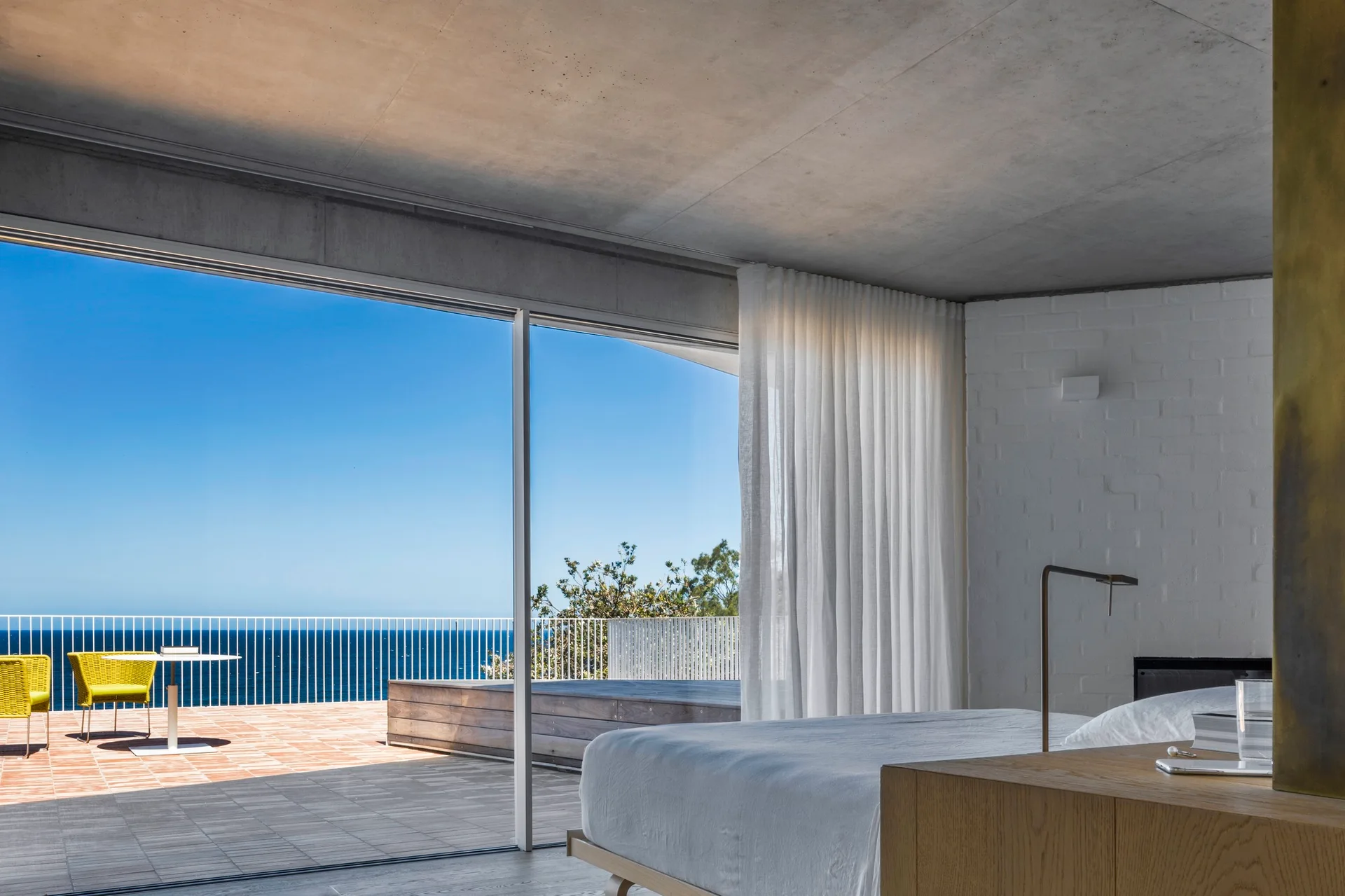 A modern bedroom with a concrete ceiling, painted brick wall, and sliding glass doors that open to a sunny terrace overlooking the ocean.