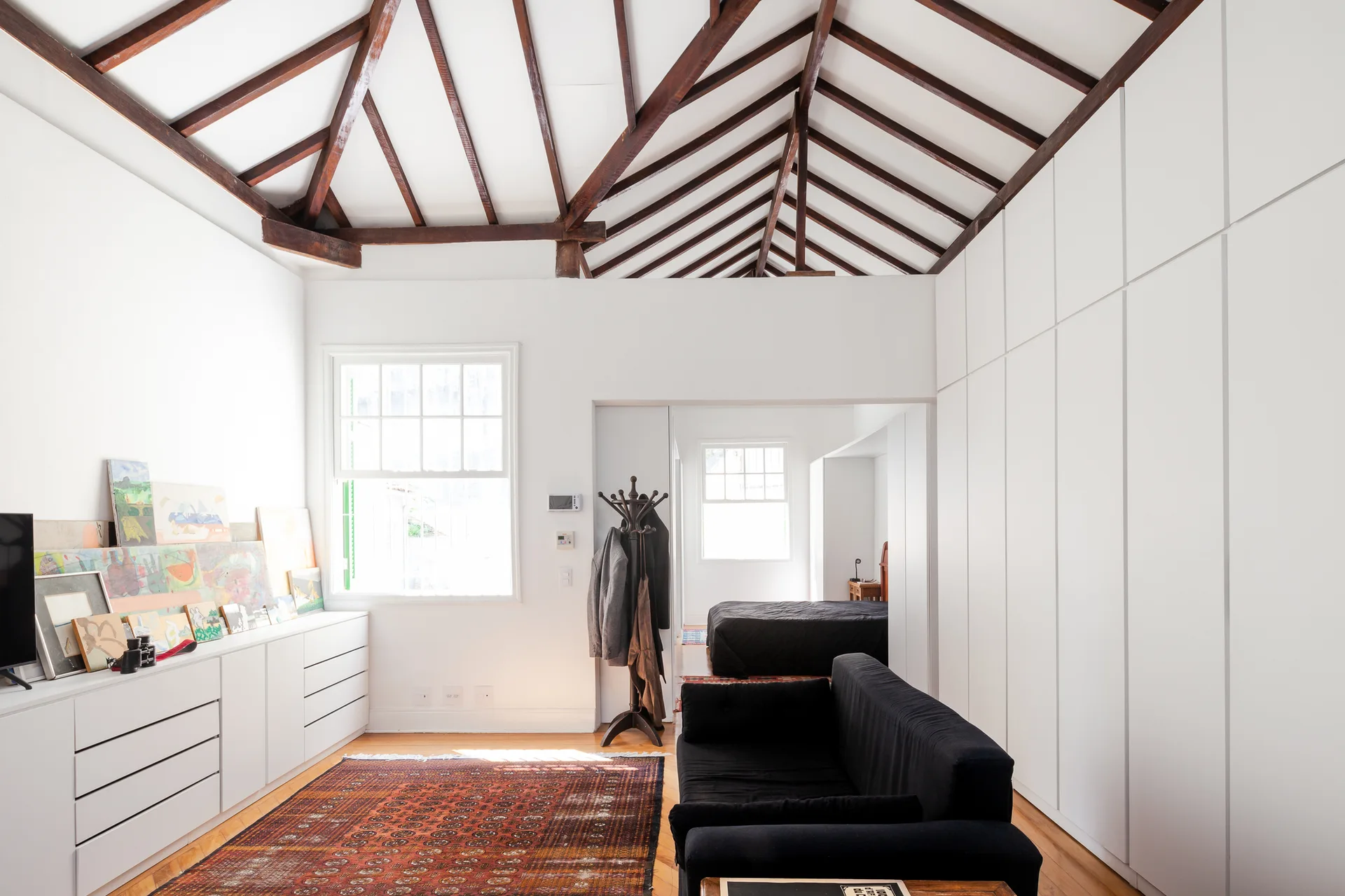 A bright living room features a black sofa, a patterned rug, white built-in storage, and a vaulted ceiling with exposed dark wood beams.