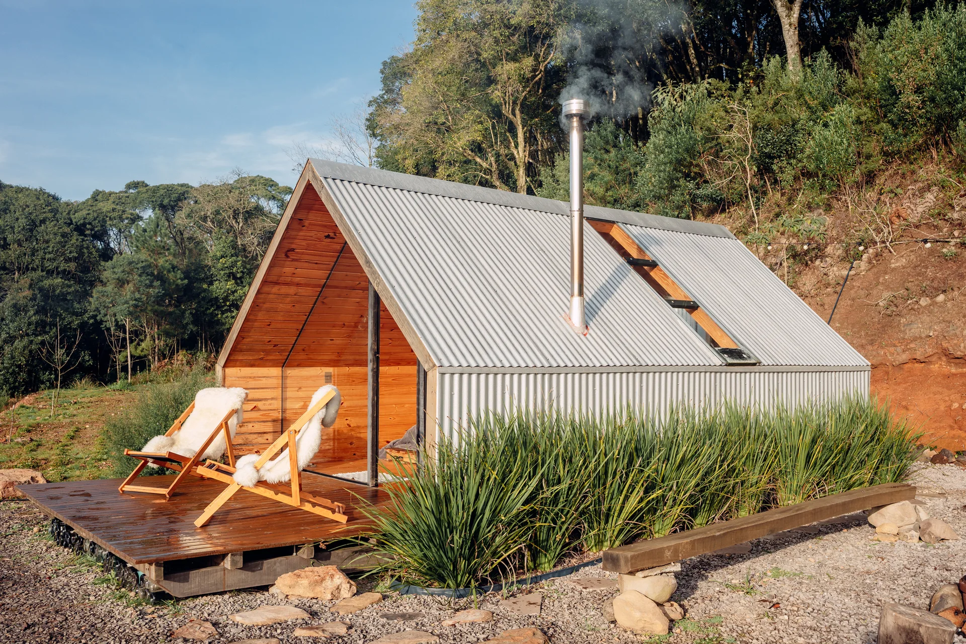 A small A-frame cabin features corrugated metal siding, a smoking chimney, and a wooden deck with two sheepskin-lined lounge chairs in a forest.