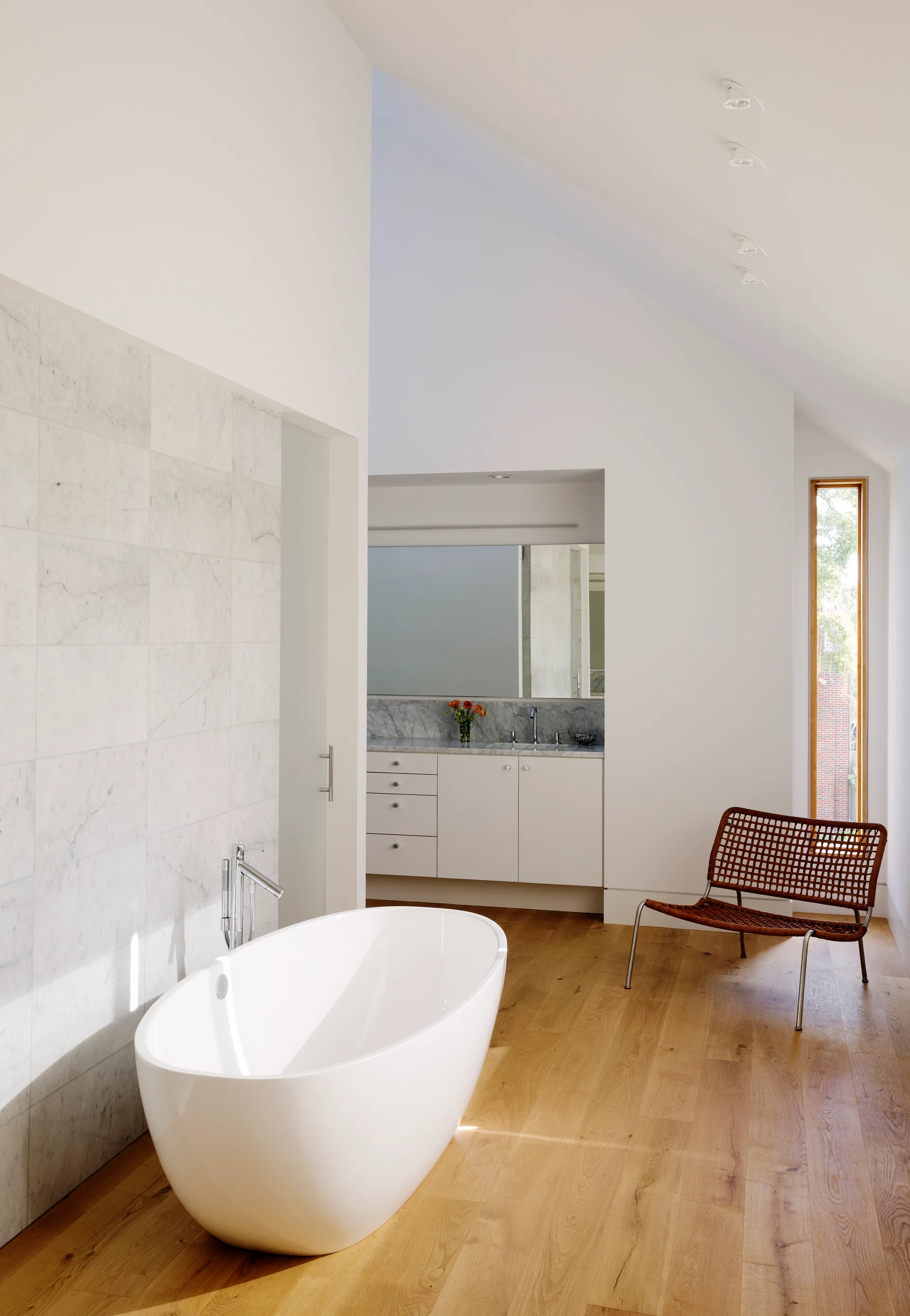 A modern bathroom features a white freestanding bathtub on wood flooring, a marble-tiled wall, and a woven lounge chair under a vaulted ceiling.