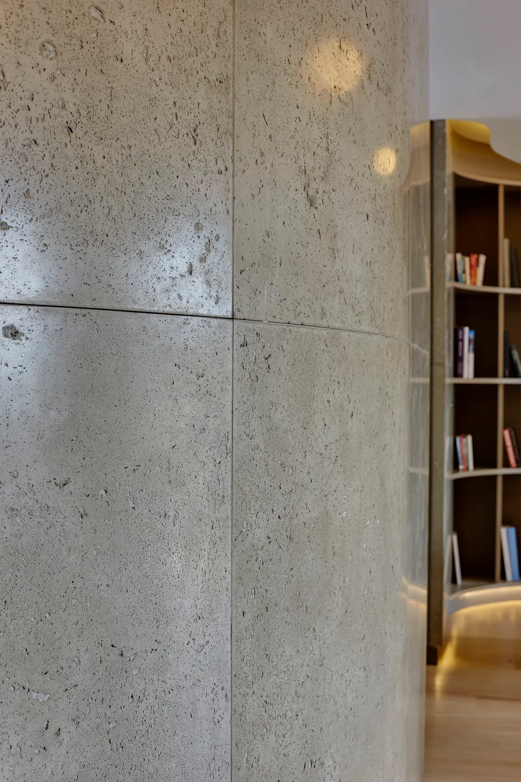 A close-up of textured greige stone panels on a curved wall next to a recessed curved wooden bookcase with books and warm indirect lighting.