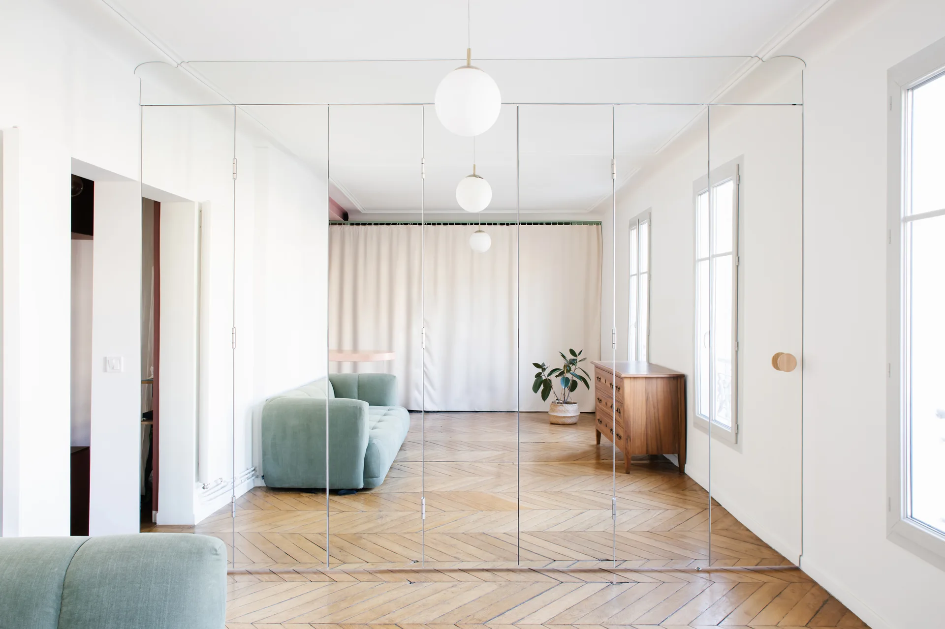 A living room features a floor-to-ceiling mirrored partition wall reflecting a sage green sofa, a wooden dresser, and herringbone parquet flooring.