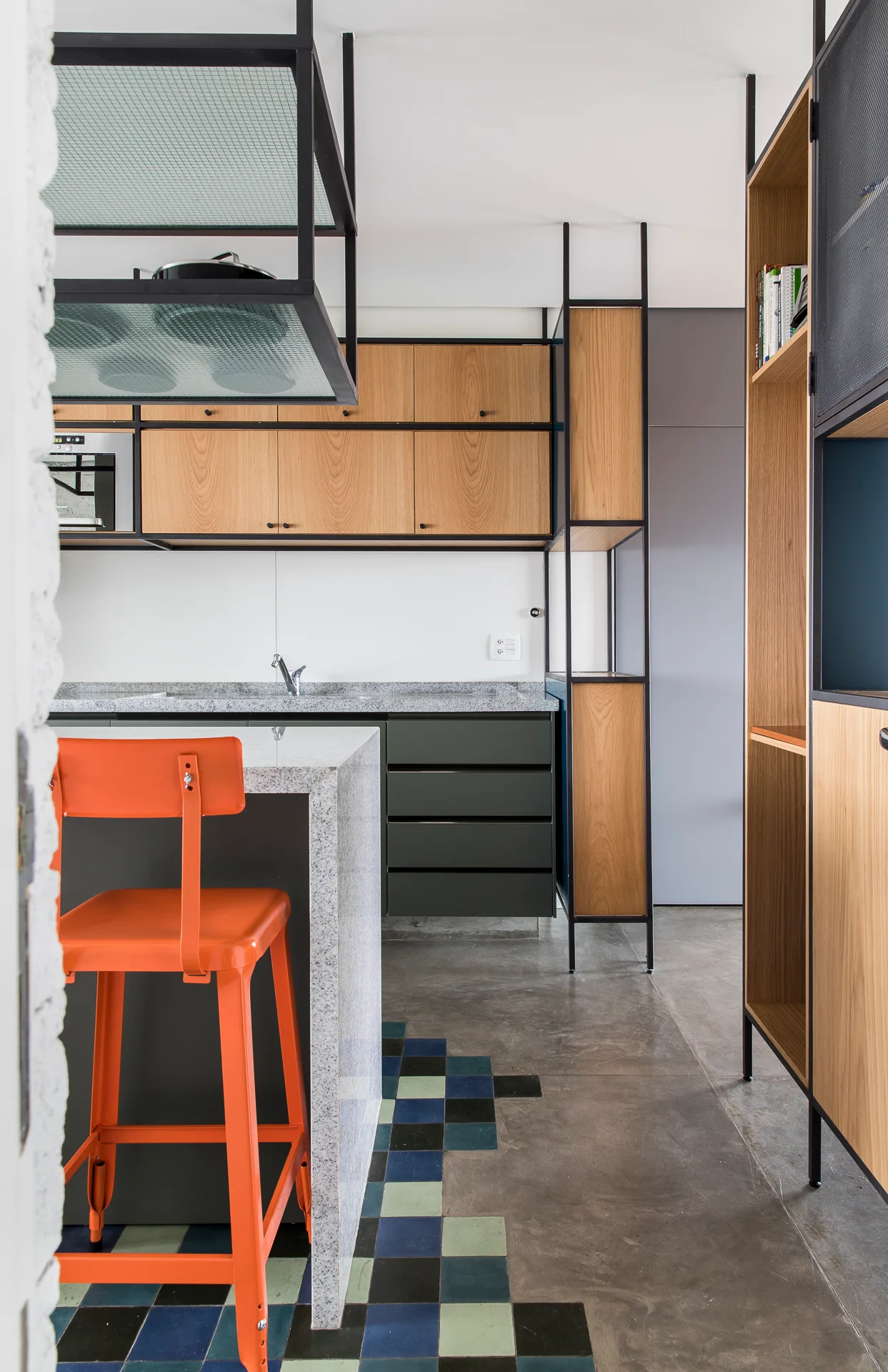 An industrial kitchen featuring wood cabinets, a speckled stone island with an orange stool, and concrete flooring with a colorful tile inlay.