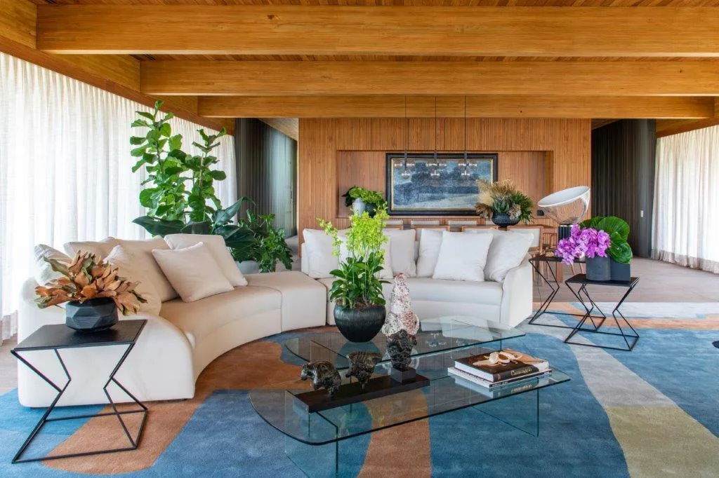 A spacious living room featuring warm wood-paneled walls, a curved cream sectional sofa, glass coffee tables, and a large abstract patterned rug.
