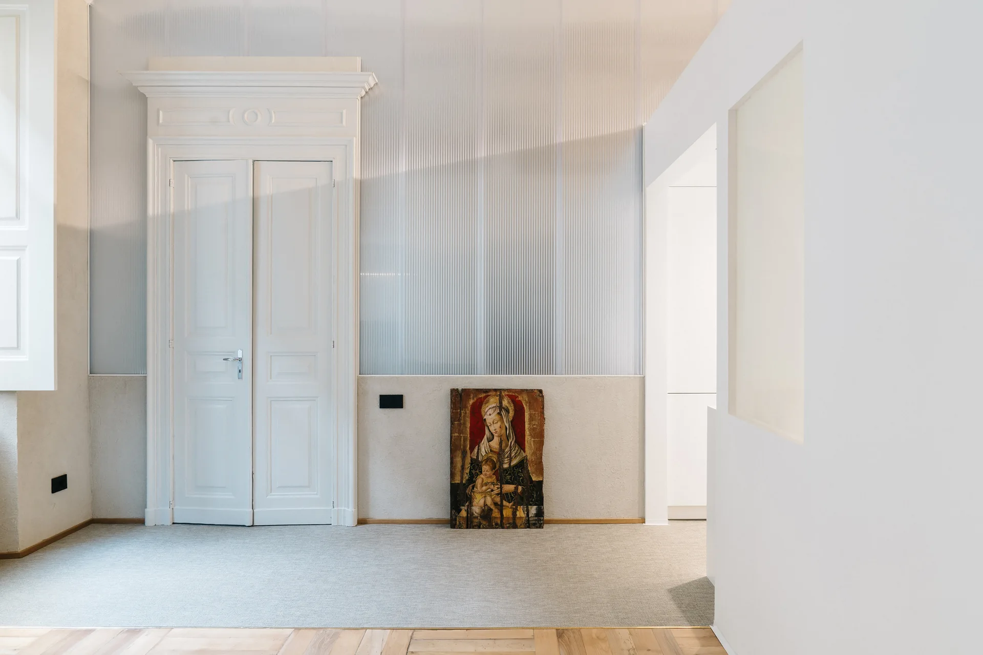 A minimalist hallway featuring a classic white double door, a translucent ribbed upper wall panel, and an art piece leaning against the floor.