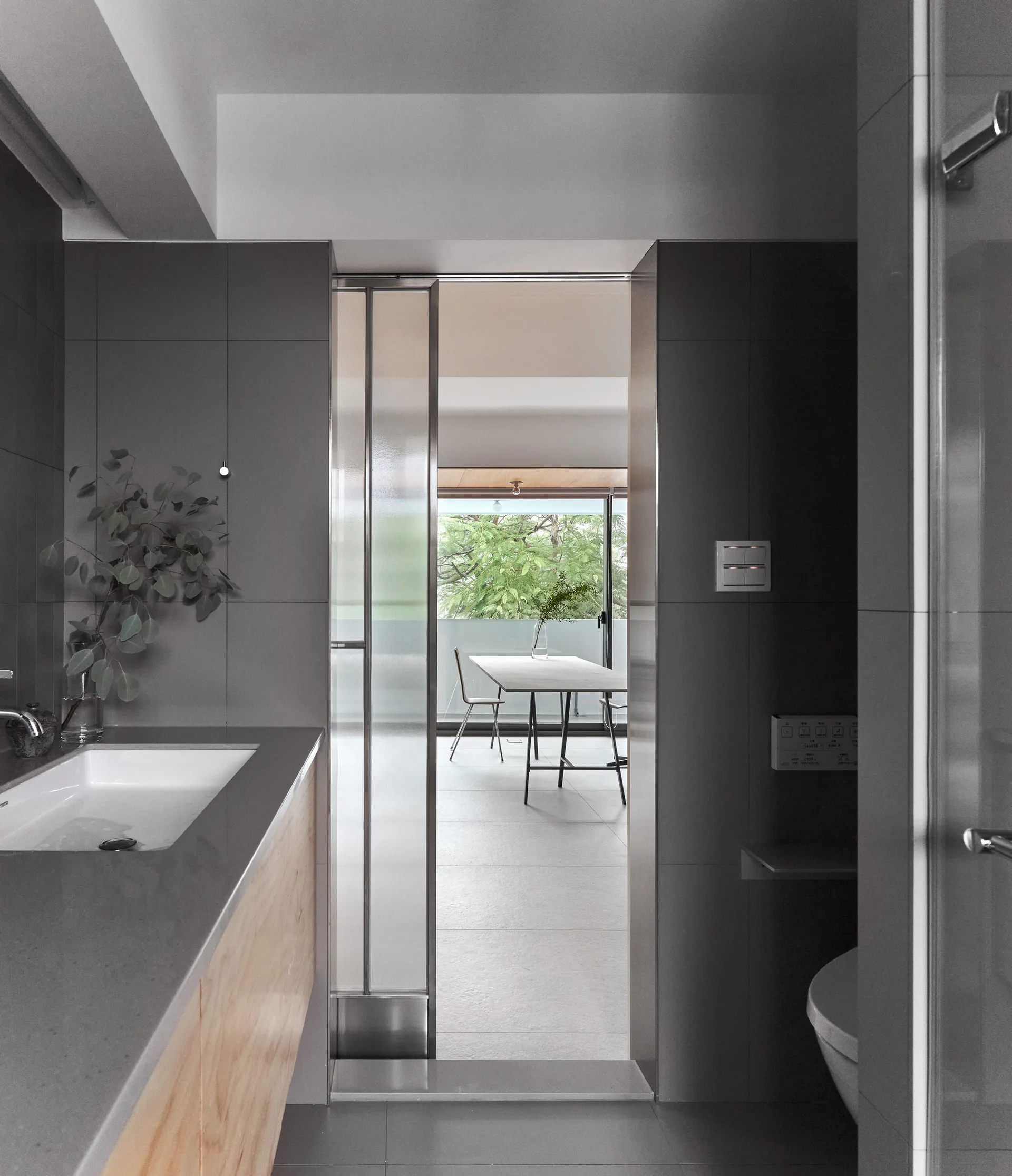 A minimalist gray tiled bathroom features a wood vanity, white rectangular sink, and a glass sliding door looking into a bright dining area.