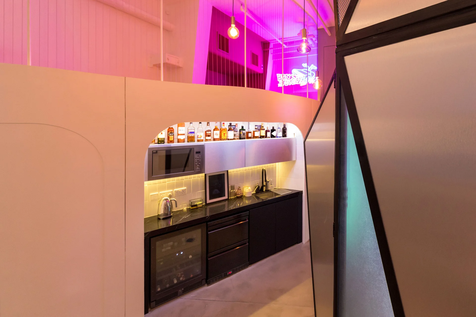 A compact black kitchenette with a marble countertop is set inside a white arched wall partition, illuminated by a pink neon glow.