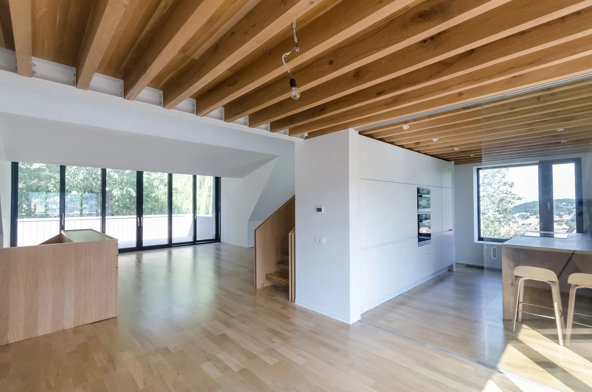 An open interior space featuring exposed wooden ceiling beams, minimalist white cabinetry, a glass partition, and light wood flooring.