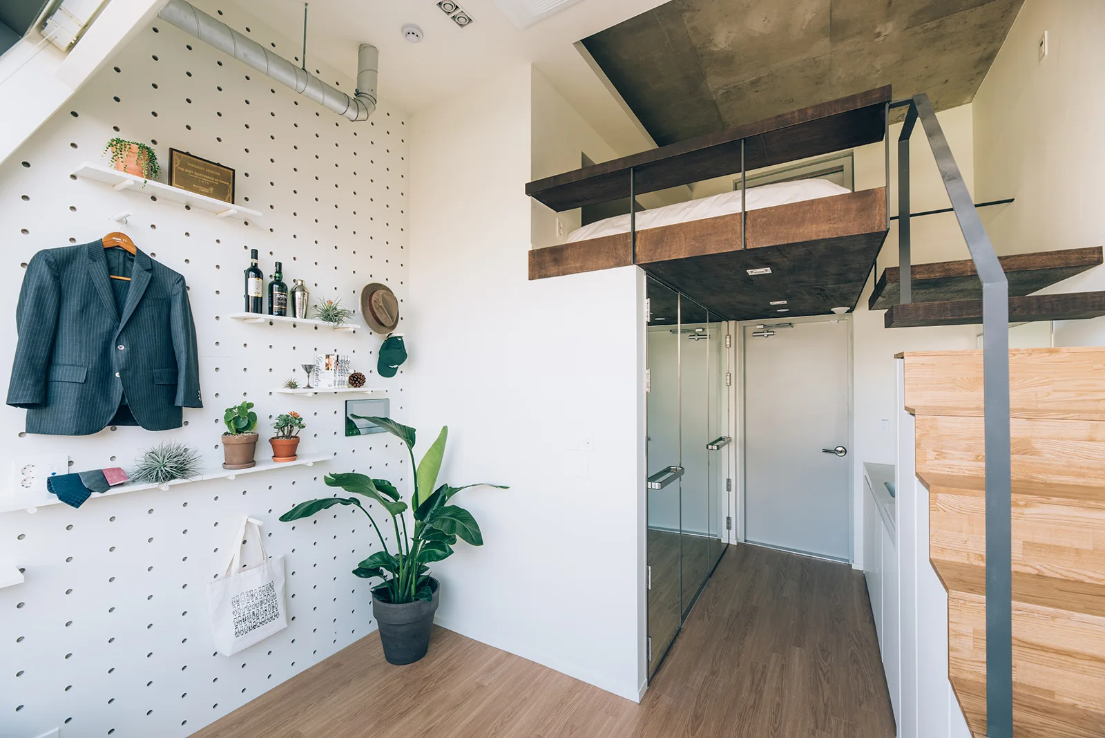 A modern studio apartment featuring a tall white pegboard storage wall, a wooden staircase, and an elevated loft bed with a concrete ceiling.