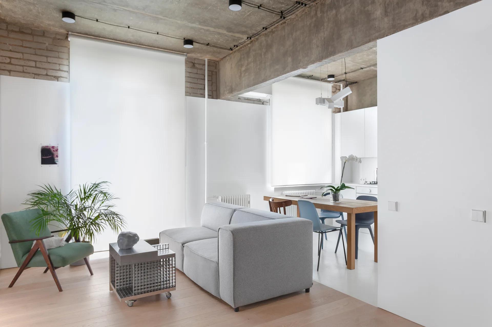 A minimalist open-plan living room features a grey sofa, a green lounge chair, and an exposed concrete ceiling looking into a dining area.