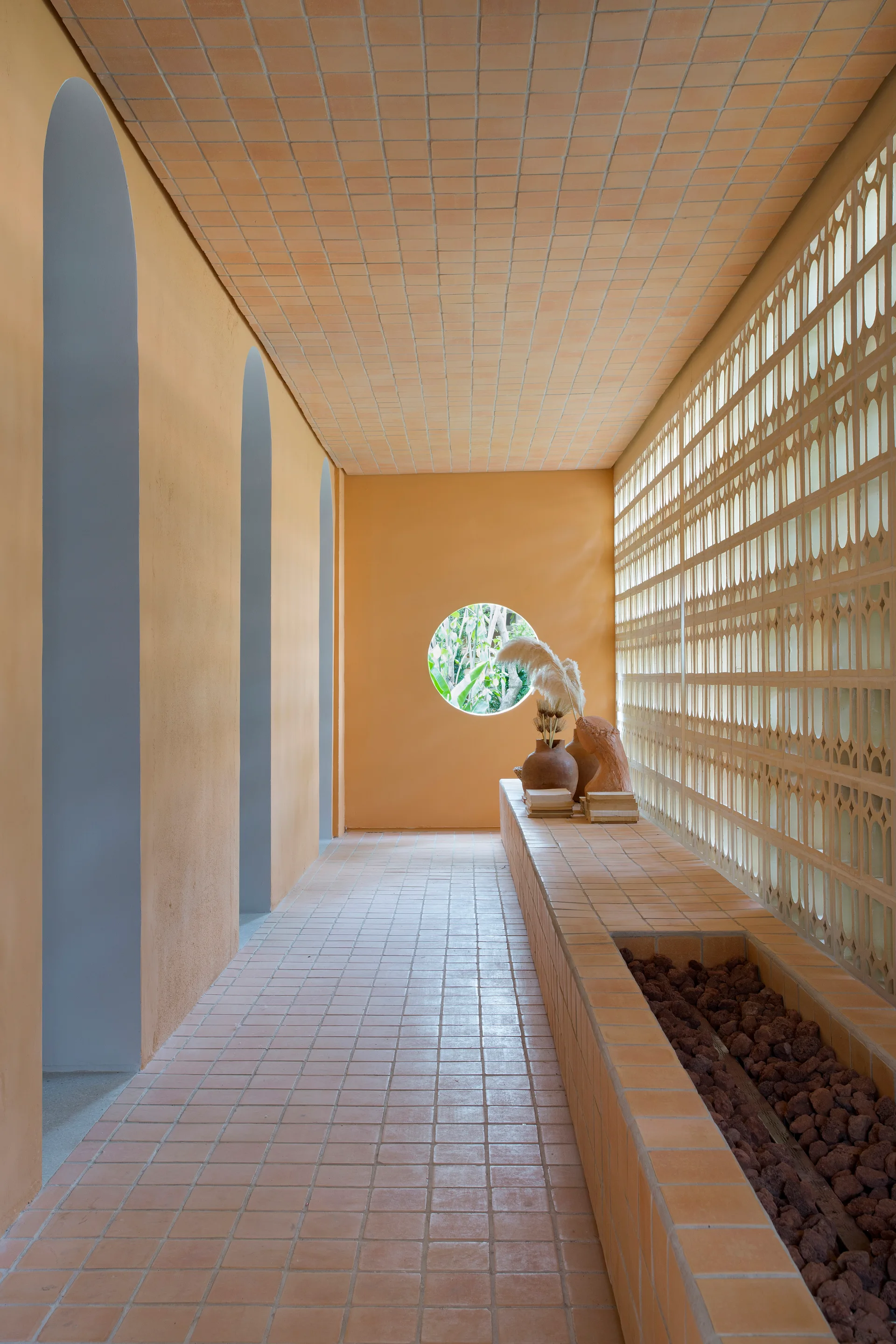 A terracotta-tiled hallway features arched wall openings on the left, a patterned breeze block screen on the right, and a circular window at the far end.