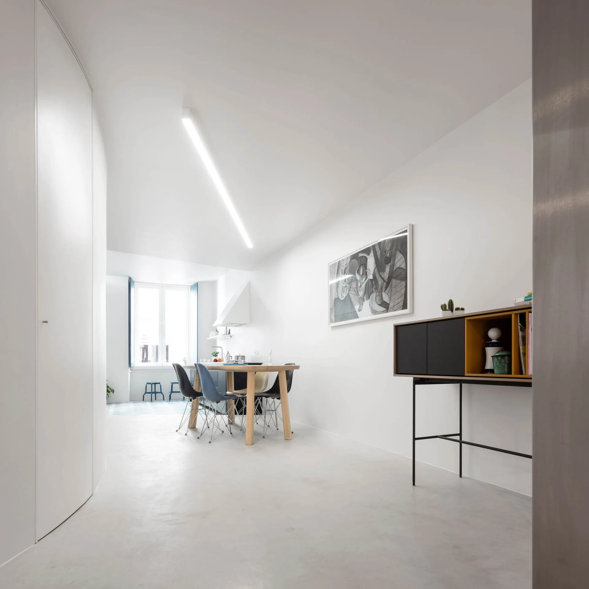A minimalist dining room features a wooden table with molded chairs, a sleek black and yellow sideboard, and a long linear light on the ceiling.