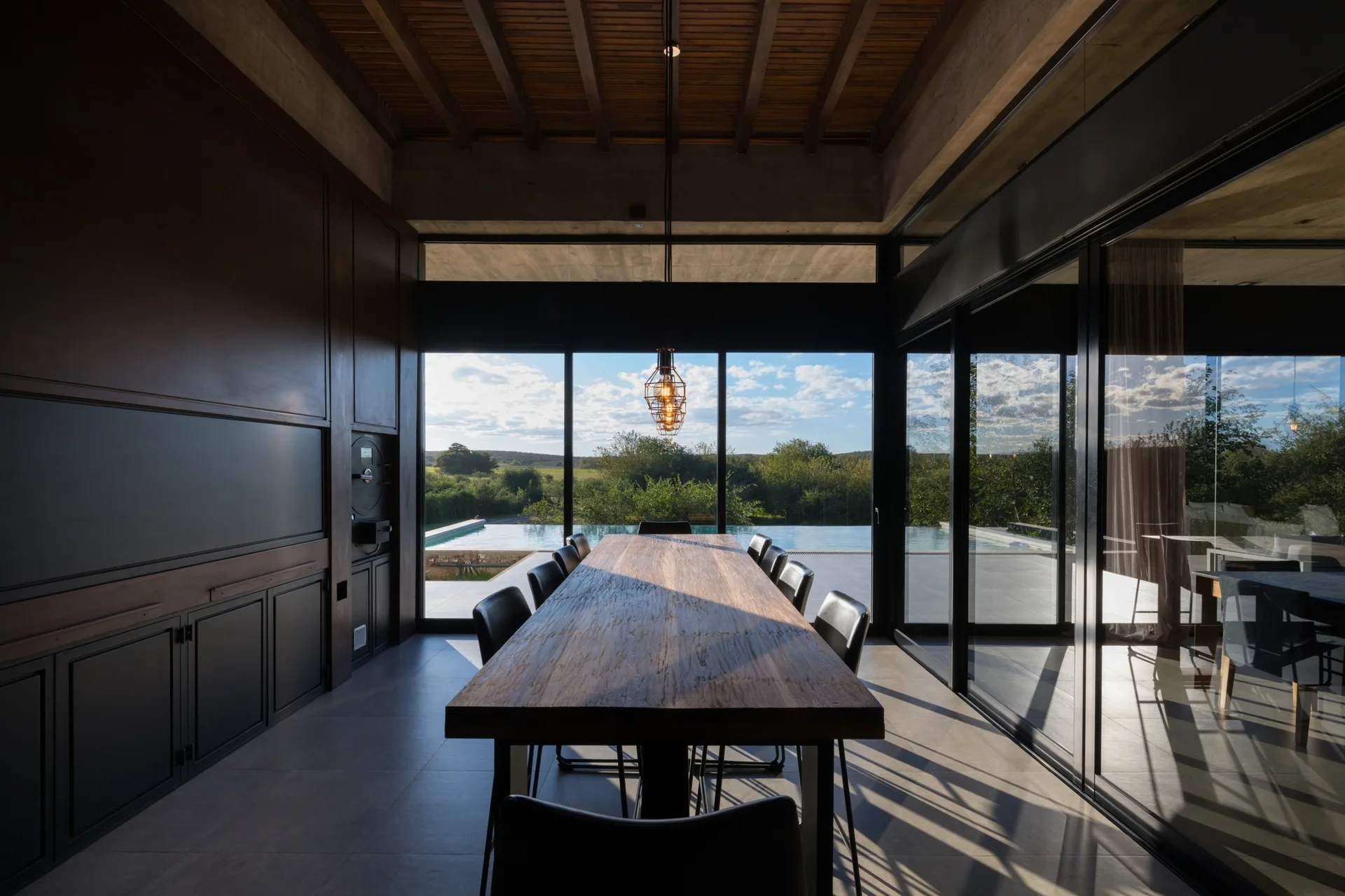 A modern dining room features a long wood table, dark wall paneling, and floor-to-ceiling glass walls looking out to a swimming pool.