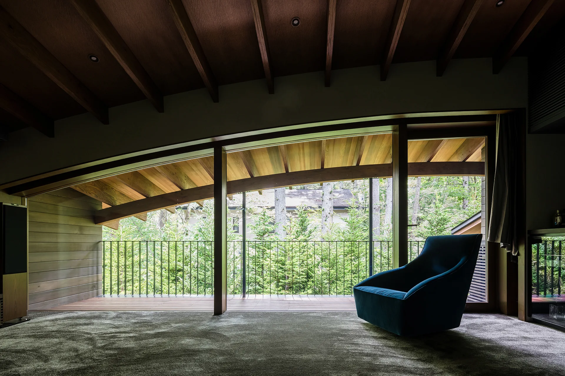 A blue armchair sits on a grey carpeted floor in a modern room with a sloped wood ceiling and a glass wall overlooking green trees.