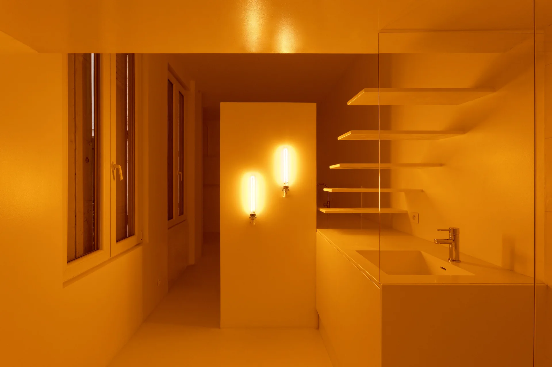 A minimalist bathroom interior bathed in warm amber light, featuring a clean white vanity, floating shelves, and two vertical wall lamps.