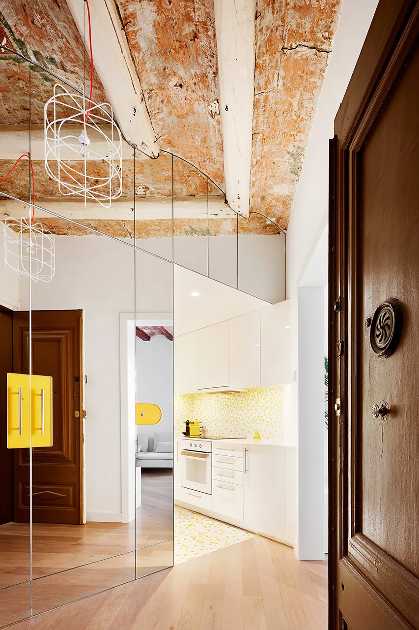 A modern white kitchen with a yellow mosaic backsplash is reflected in large mirrored wall panels beneath an exposed vaulted brick ceiling.
