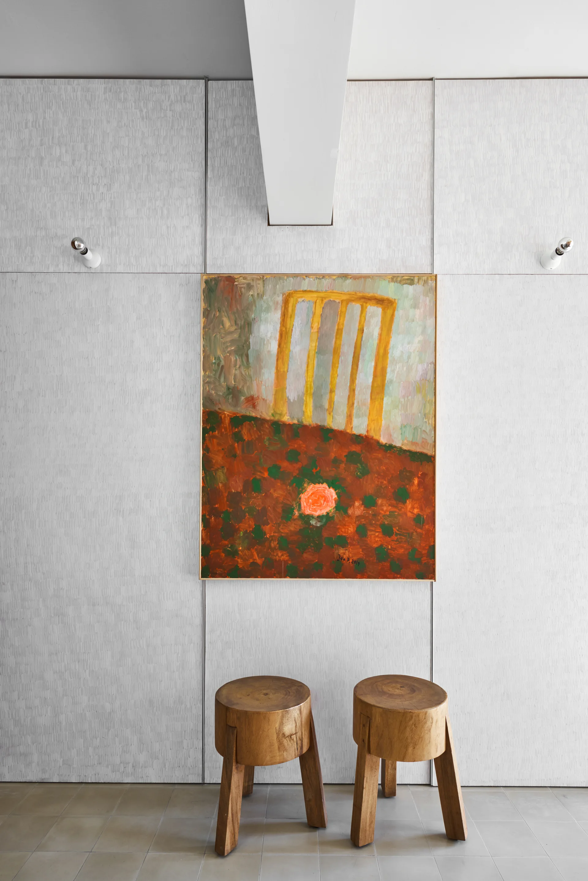 Two rustic wooden stools stand on a tiled floor below a colorful painting mounted on a textured light grey paneled wall.