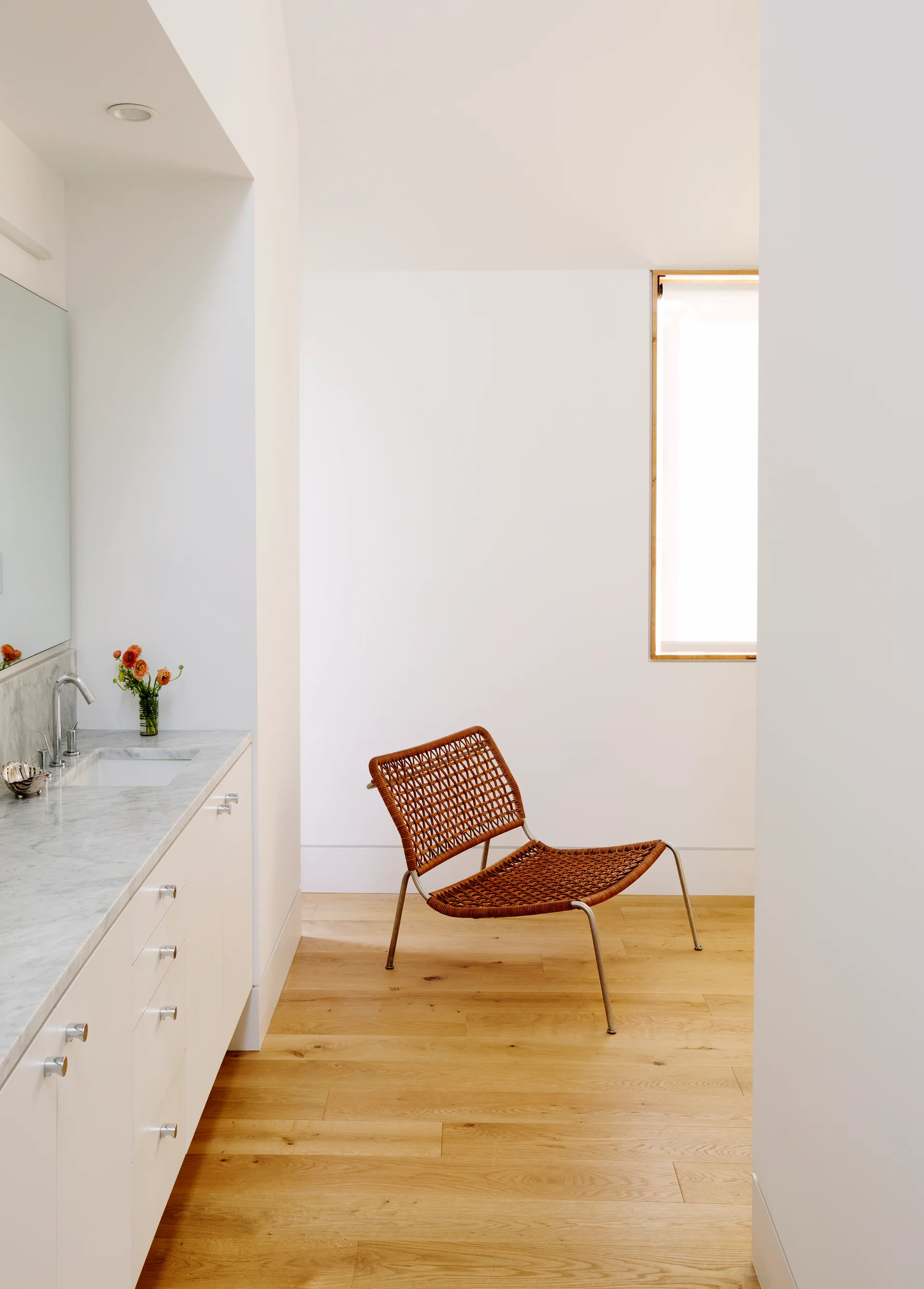 A bright minimalist bathroom features a white marble vanity next to a light wood floor hallway where a brown woven lounge chair sits near a window.