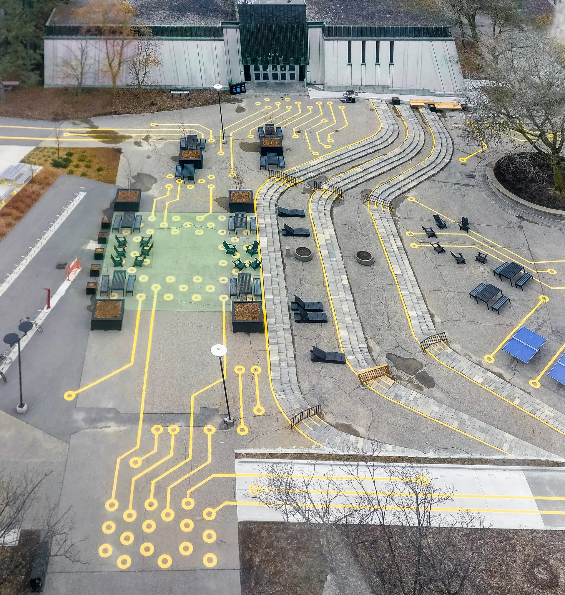 An aerial view of a paved outdoor courtyard decorated with bright yellow painted circuit board patterns, black benches, and tiered stone pathways.