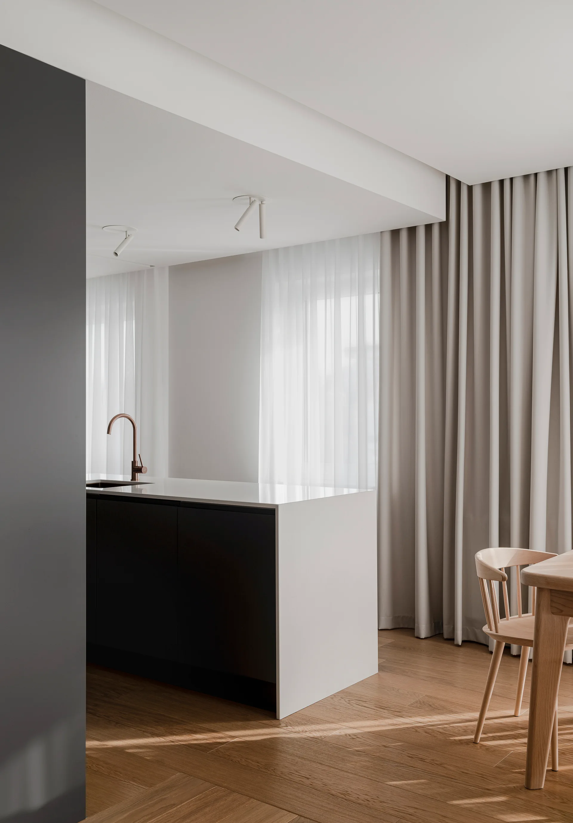 A minimalist kitchen island with a dark base, white waterfall countertop, and copper faucet stands near a dining table and floor-to-ceiling curtains.