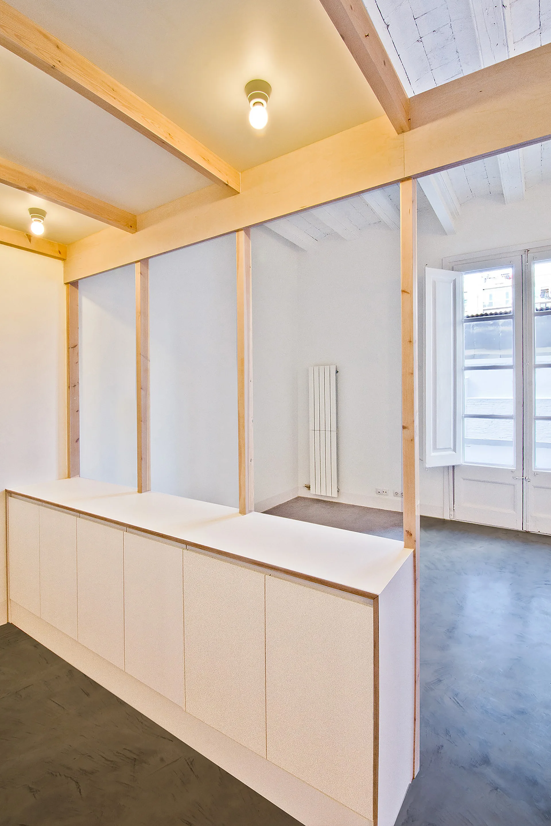 A minimalist room featuring white built-in storage cabinets under a raw timber frame partition, with grey concrete floors and white walls.