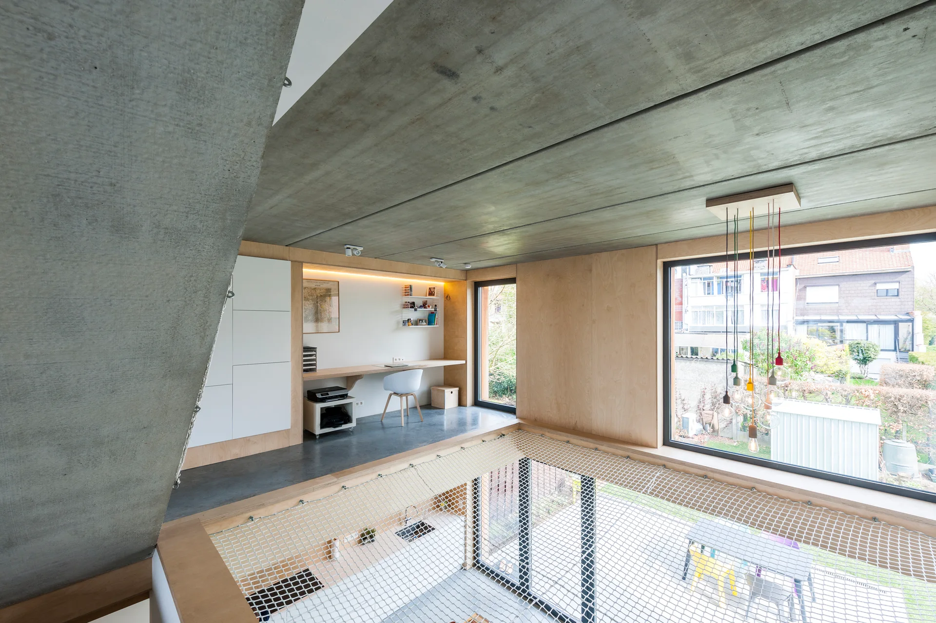 A modern mezzanine home office with a built-in wooden desk, plywood wall panels, an exposed concrete ceiling, and a safety net guardrail.