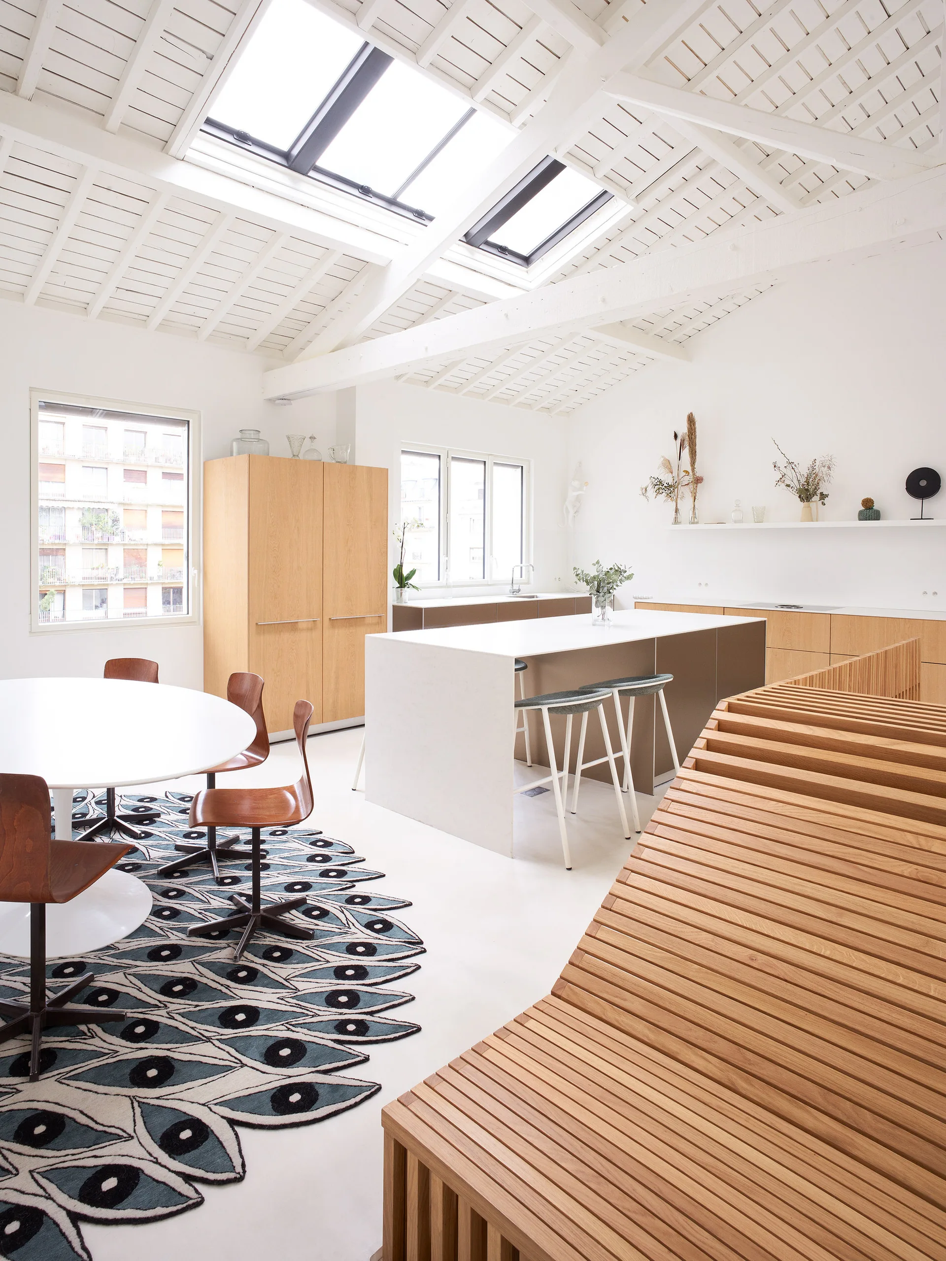 A white open-plan kitchen featuring a large skylight, wood cabinetry, a central island, and a dining table resting on an eye-patterned rug.