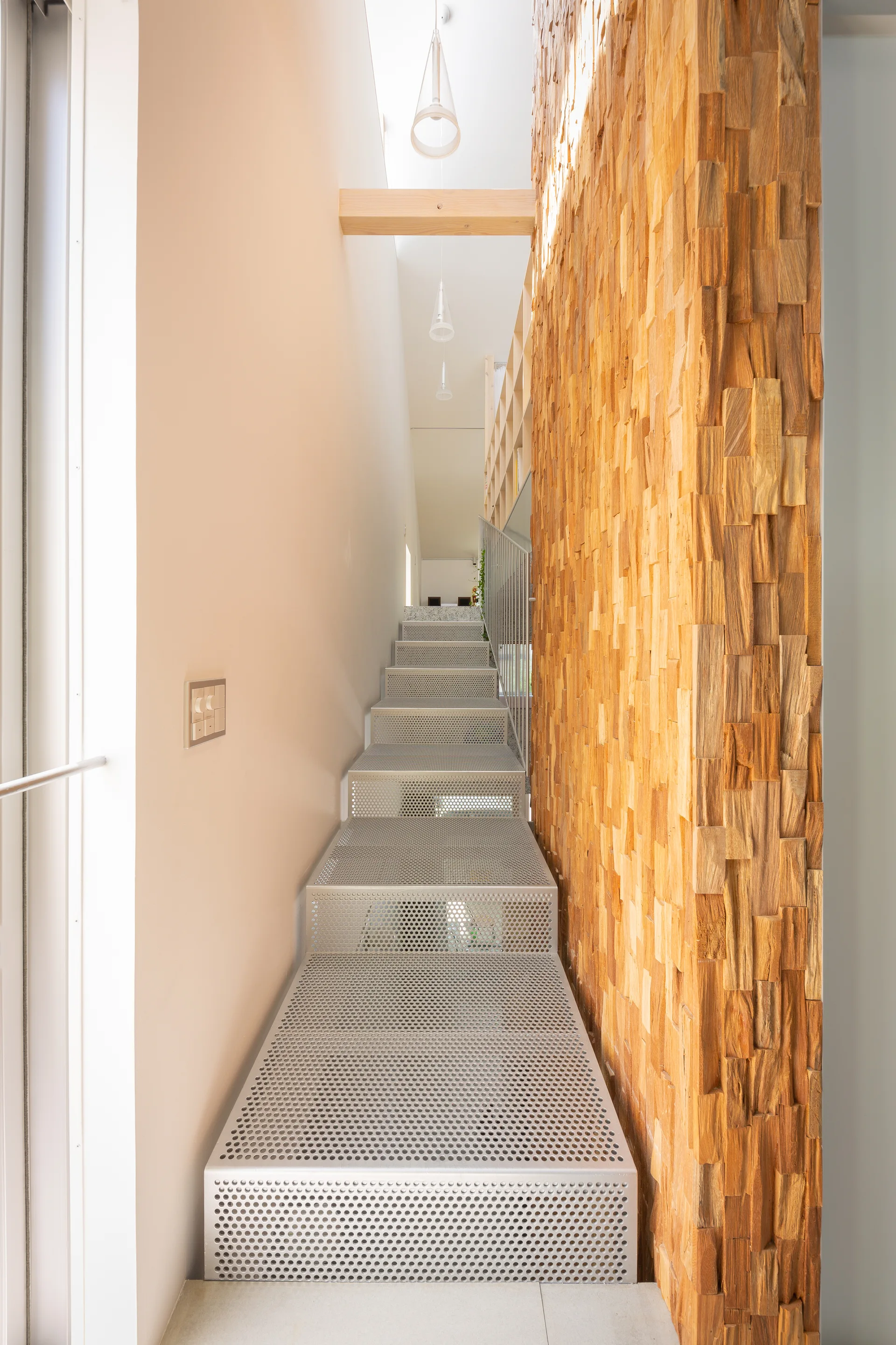 A narrow stairwell featuring perforated metal steps alongside a tall textured wood block feature wall under pendant lighting.