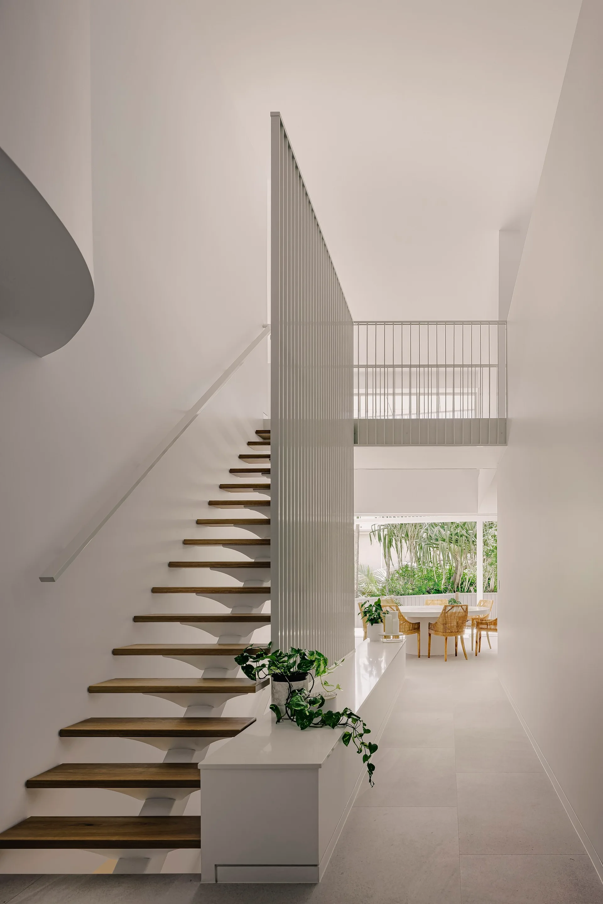 A minimalist white staircase features open wooden treads and a tall vertical slat divider, looking toward a bright dining area.