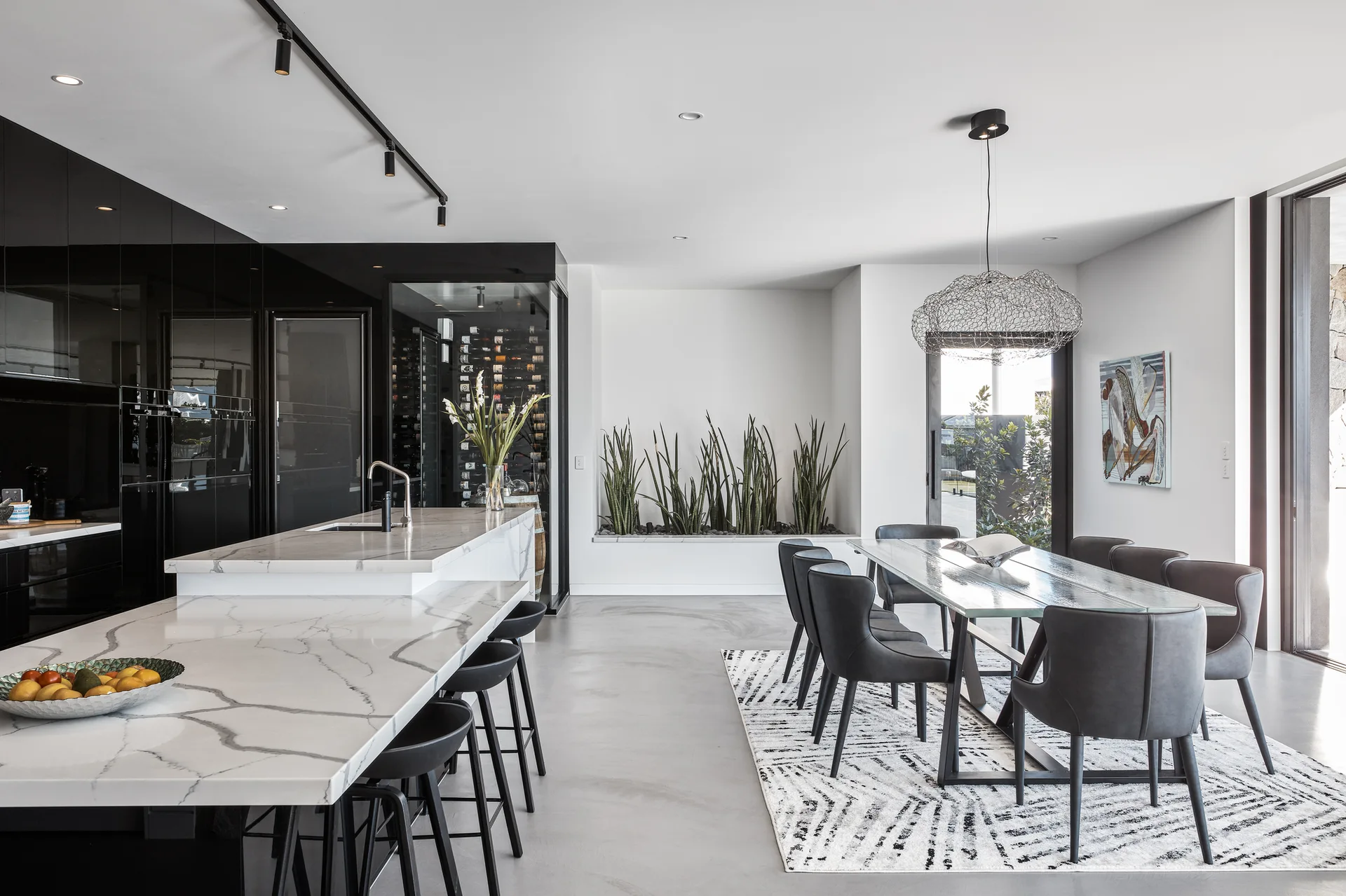 A modern open-plan kitchen and dining area featuring a white marbled island, reflective black cabinets, and a dining table over a geometric rug.