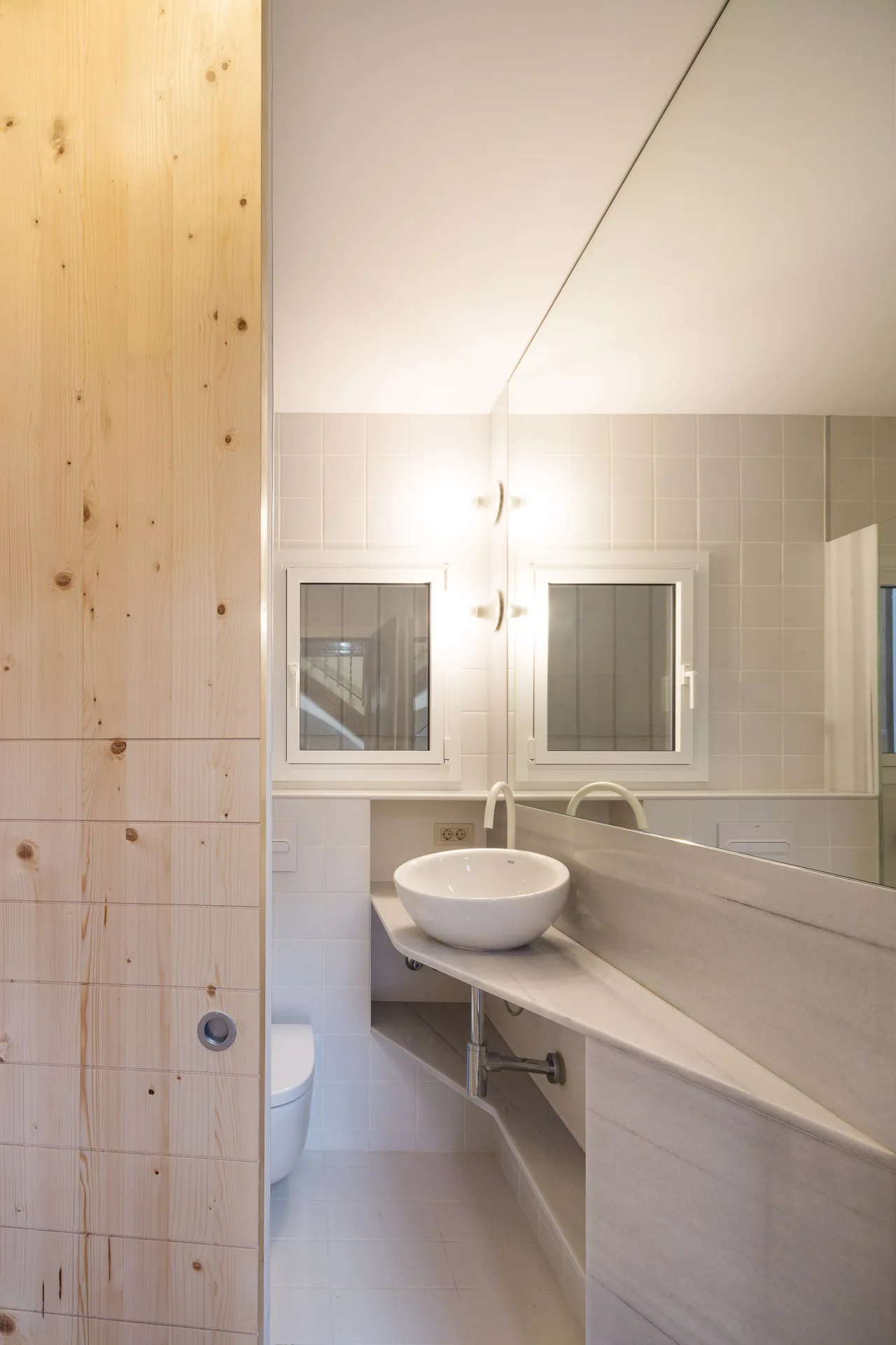 A modern bathroom with a white vessel sink on a stone shelf, a wall-to-wall mirror, and a light wood sliding door partition.