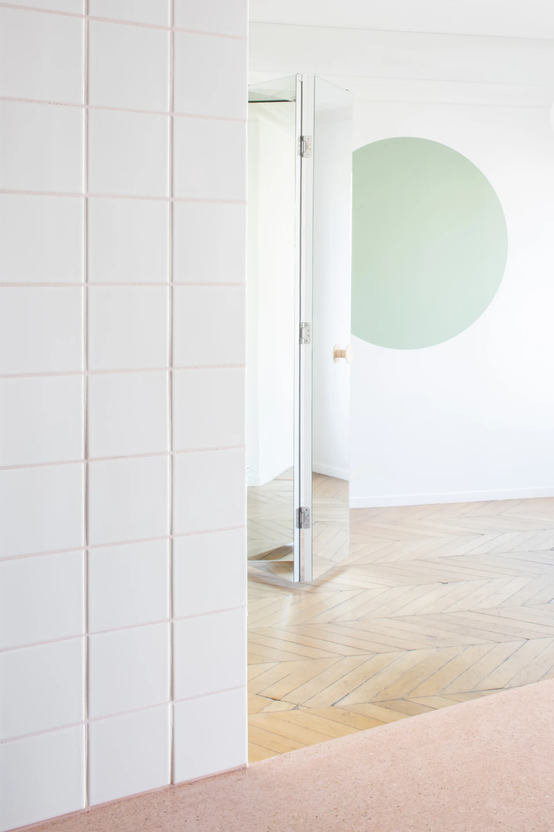 A modern interior features white grid wall tiles, a pink terrazzo floor transition, a mirrored door, and a green circular wall mural.